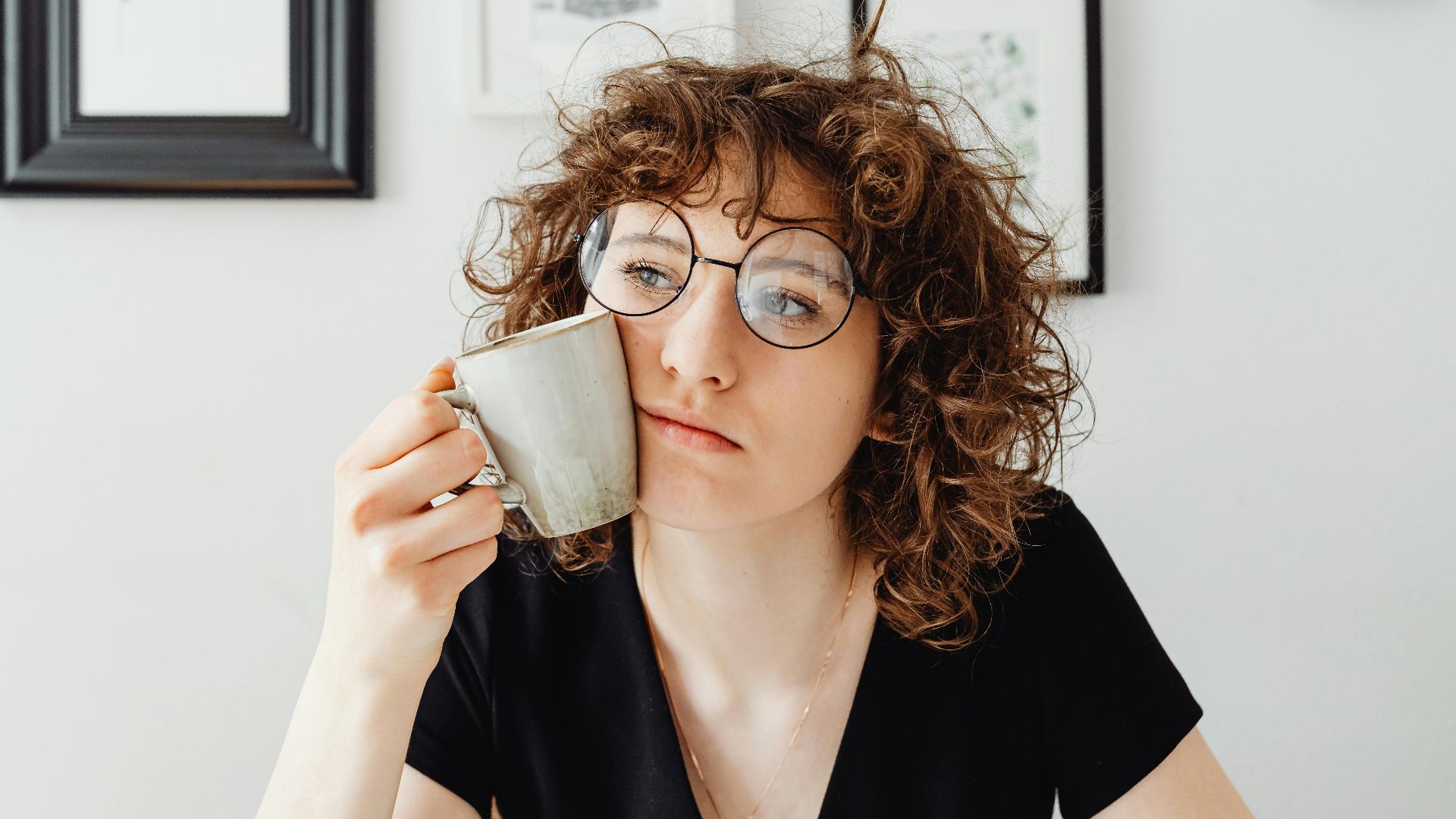 A thoughtful woman with curly hair and glasses holding a coffee mug in a modern room.