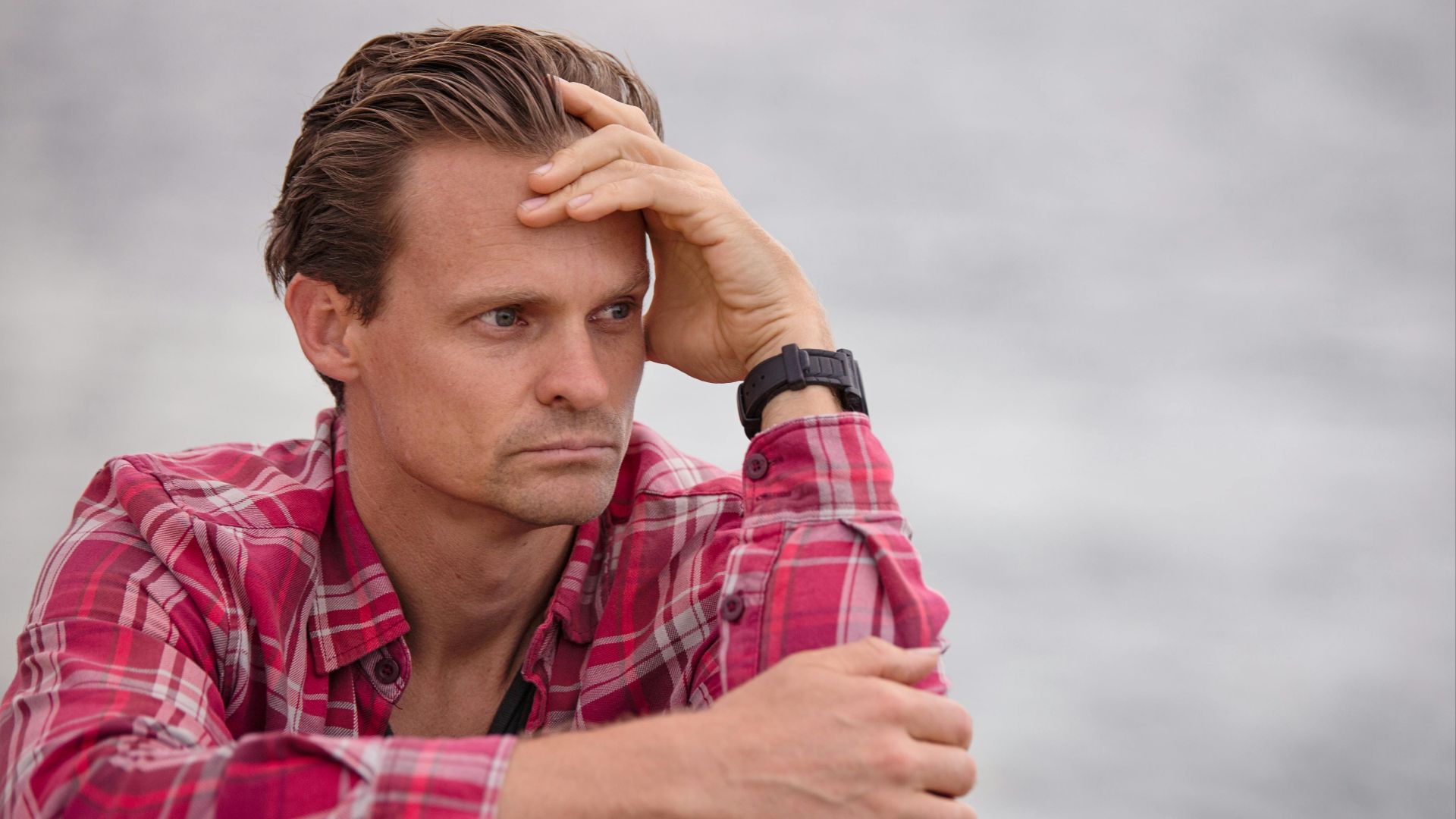 Adult male in plaid shirt sitting outdoors, deep in thought, with casual and relaxed demeanor.