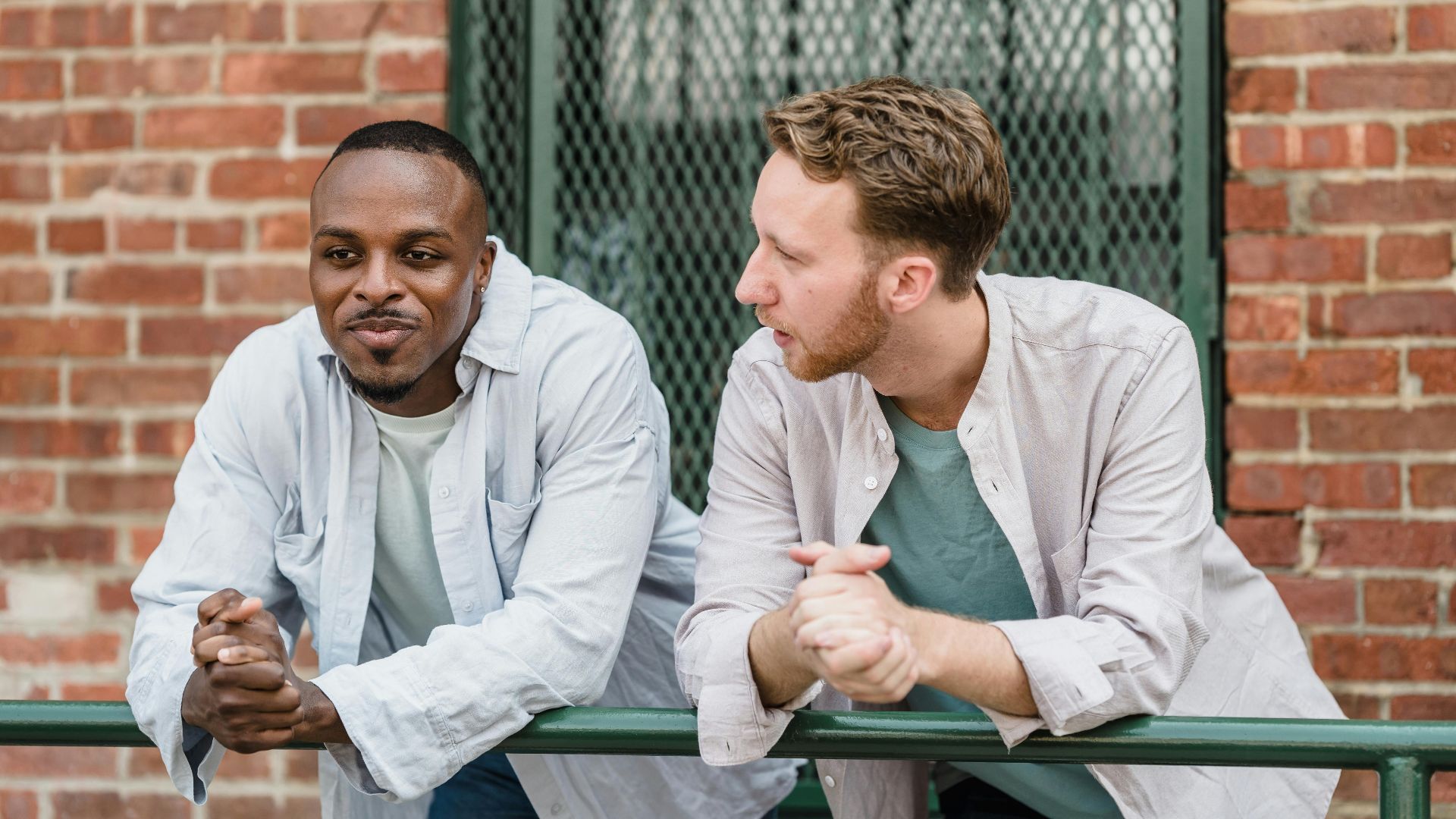 Two men having a relaxed conversation outside against a brick wall backdrop.