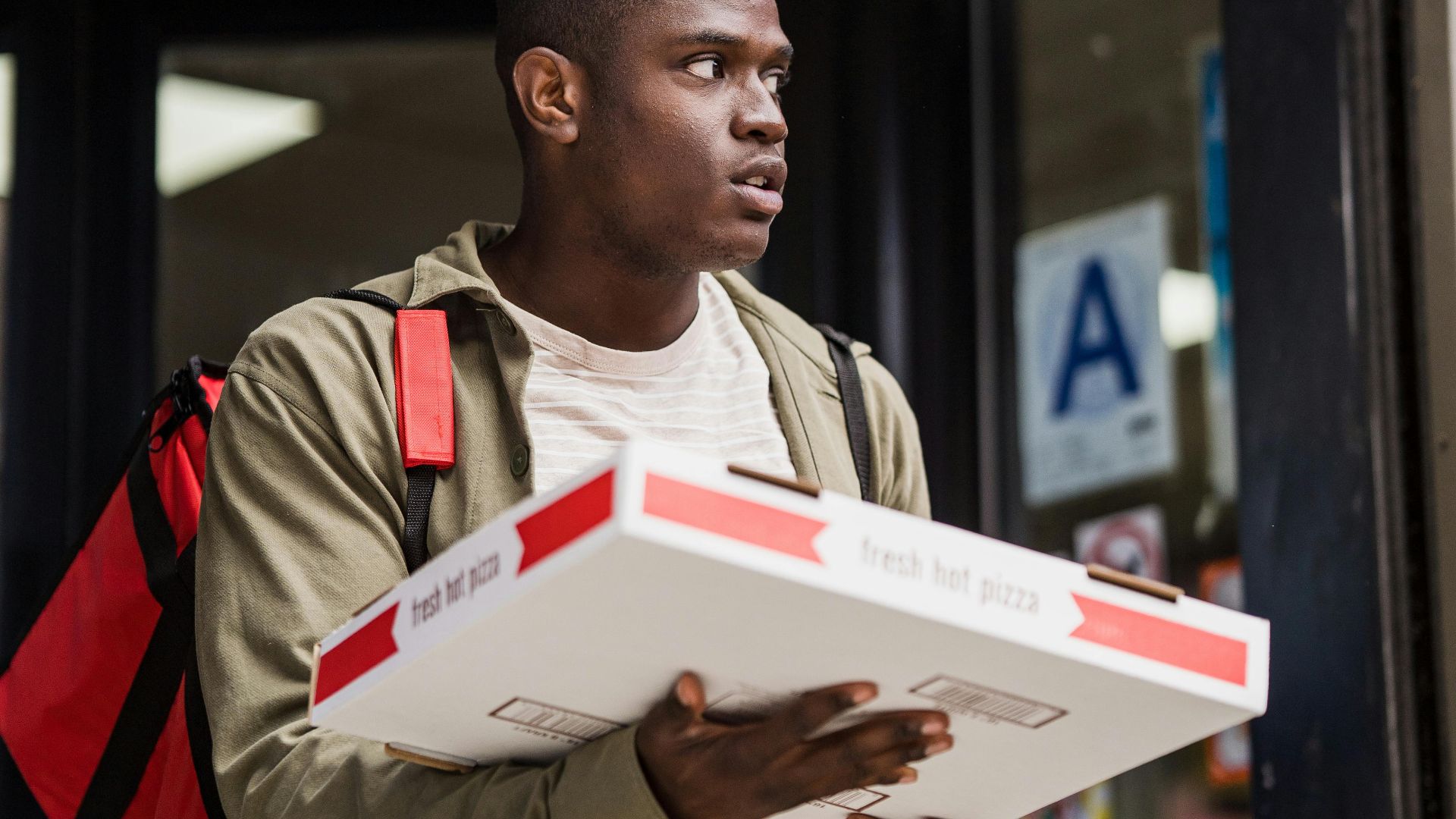Young delivery person with a thermal bag carrying a pizza box outside a restaurant.