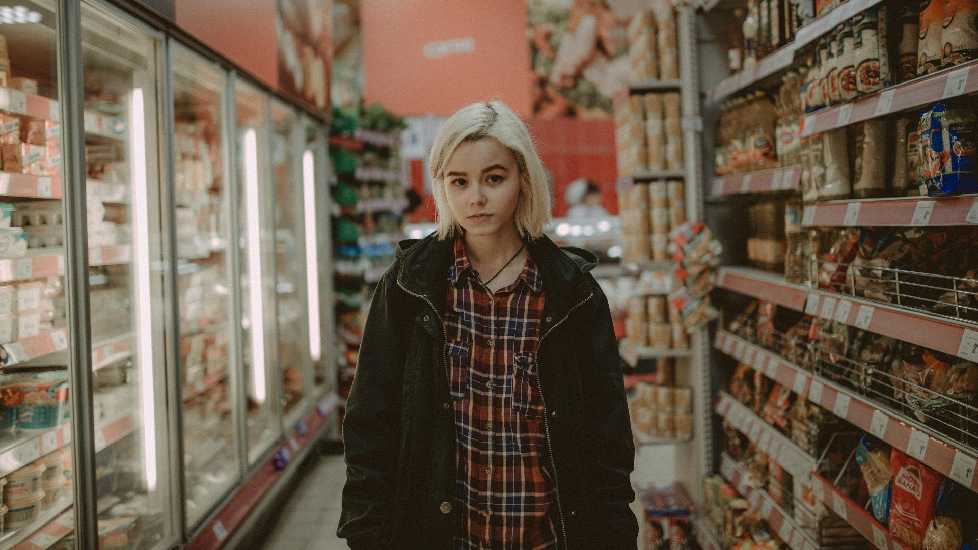 A woman with white hair stands between shelves in a supermarket aisle, showcasing a variety of products.