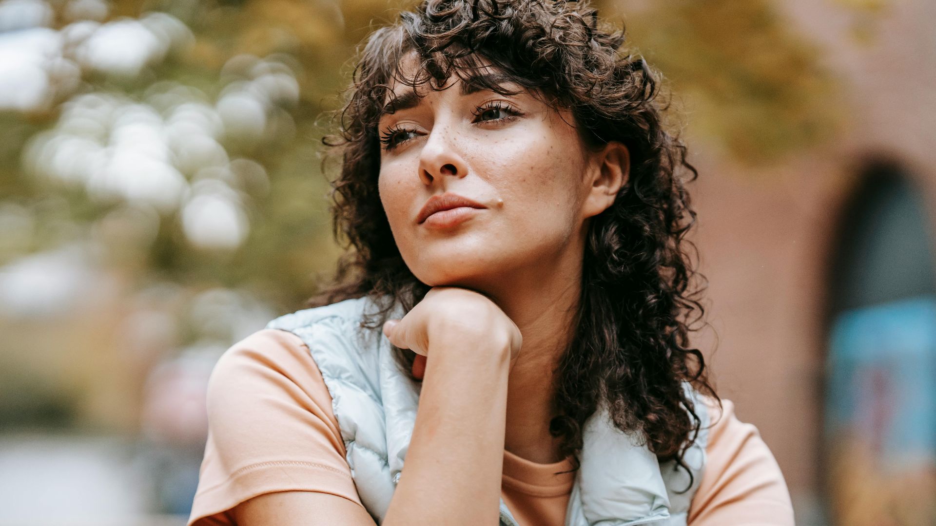 Low angle of crop pensive female with dark curly hair touching chin while looking away