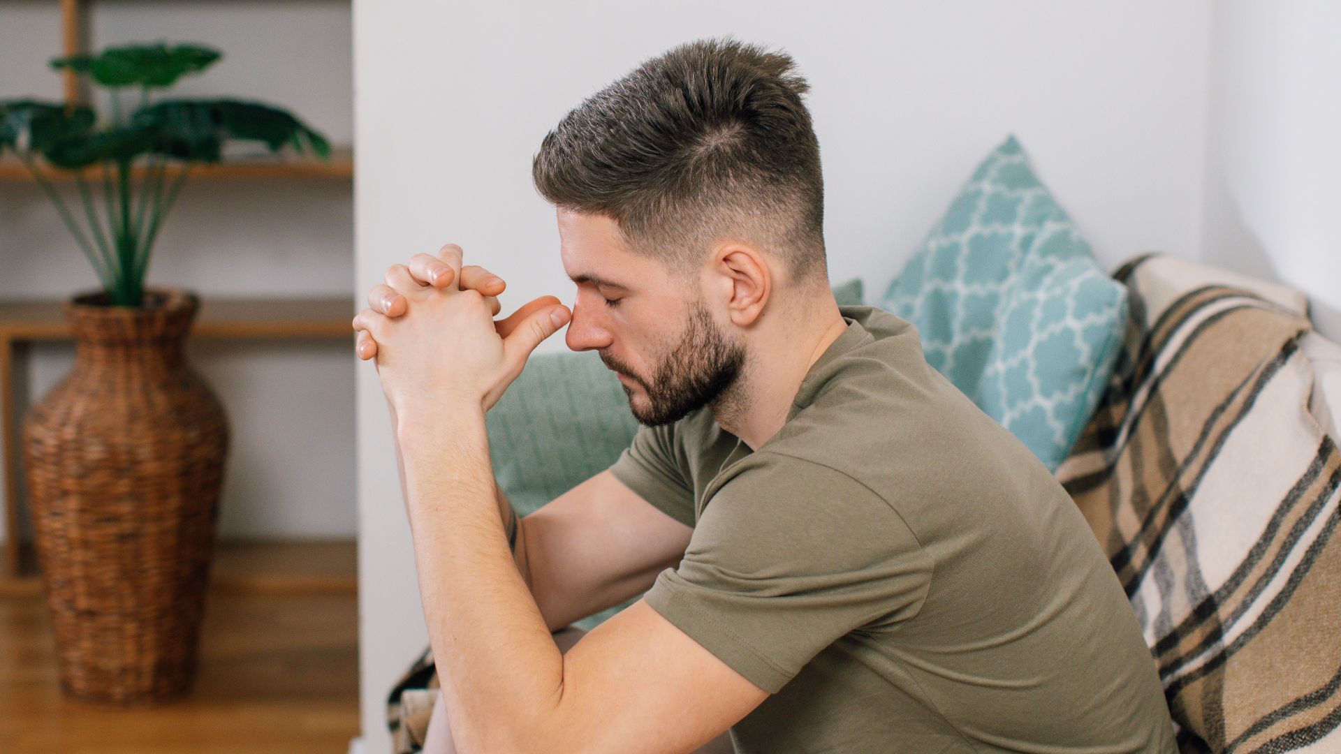A young man with clasped hands, sitting thoughtfully on a sofa in a cozy living room.