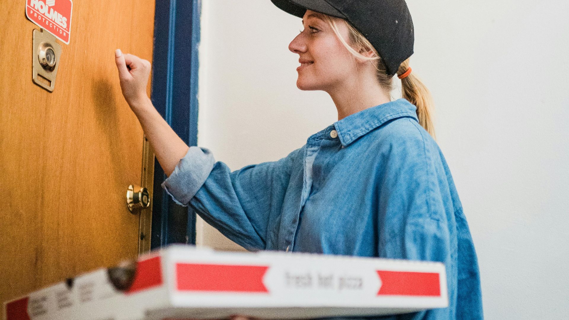 Young female pizza delivery driver knocking at an apartment door holding pizza boxes, ready for delivery.