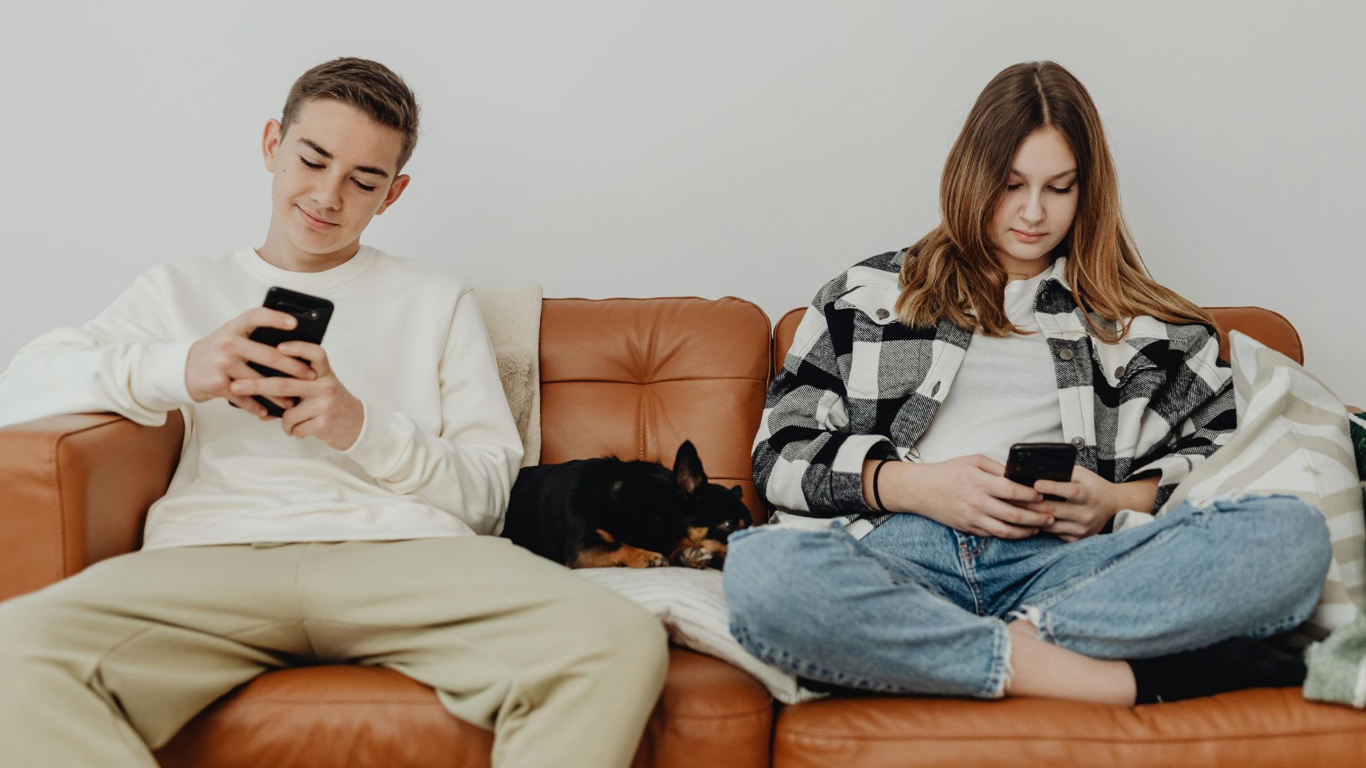 Two teenagers sitting on a sofa with a dog, using smartphones in a relaxed indoor setting.