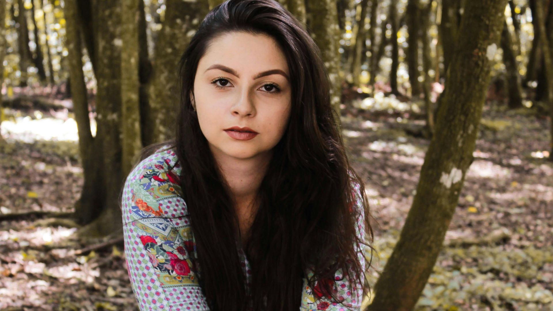 A young woman sits in a sunlit forest wearing a colorful dress, exuding tranquility.