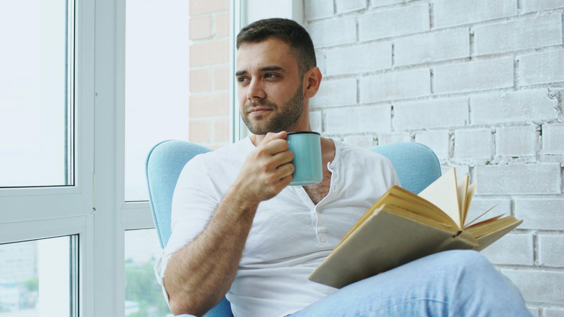 Man enjoys a quiet moment with coffee and a book in a cozy chair by the window.