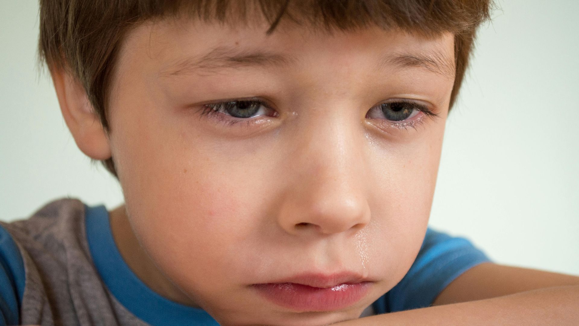 Portrait of crop upset little brown hair child with tears on face crying and resting head on arm while looking away against white background