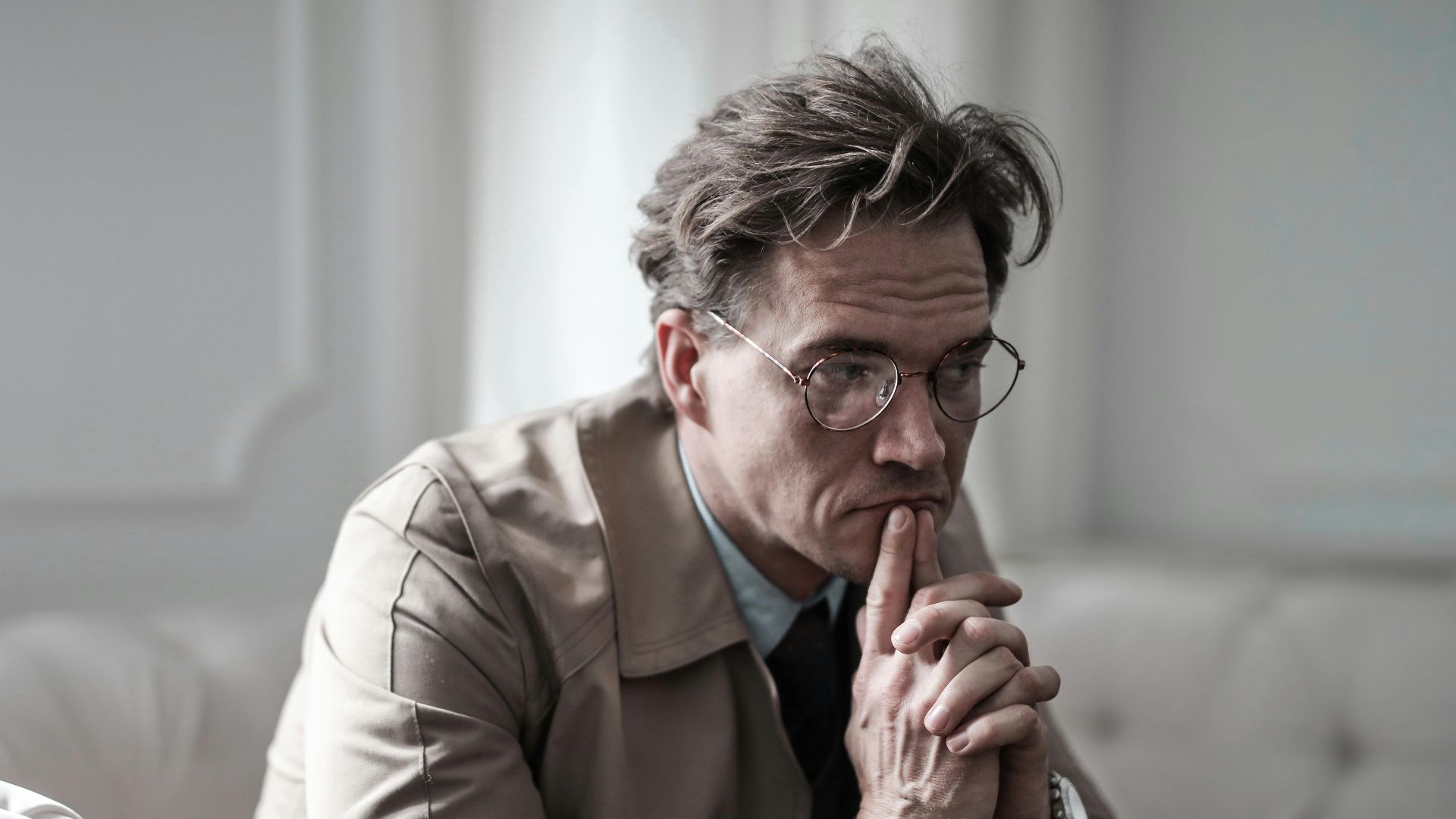 A thoughtful man wearing glasses sits on a sofa in a classic interior, lost in deep contemplation.