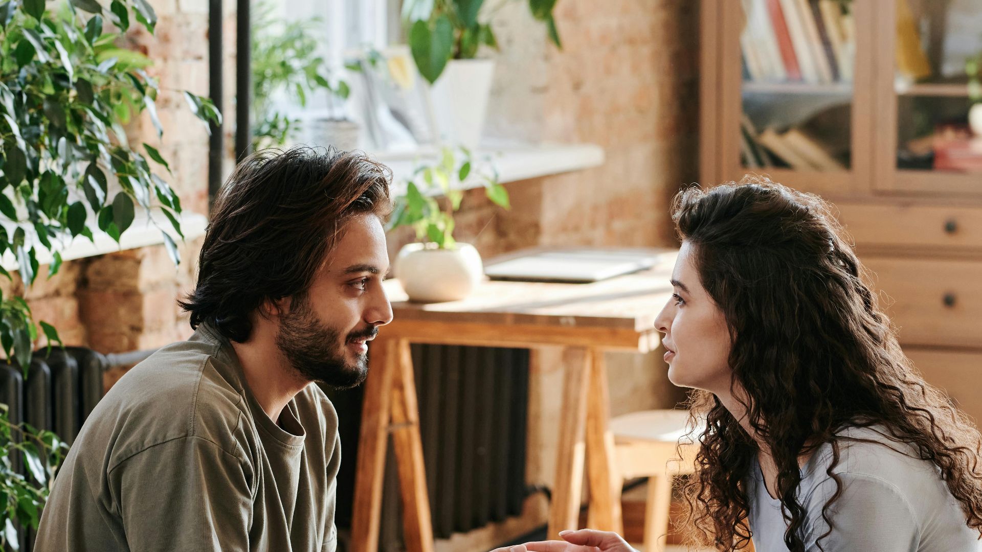 Romantic couple holding hands and talking in a cozy, plant-filled bedroom.