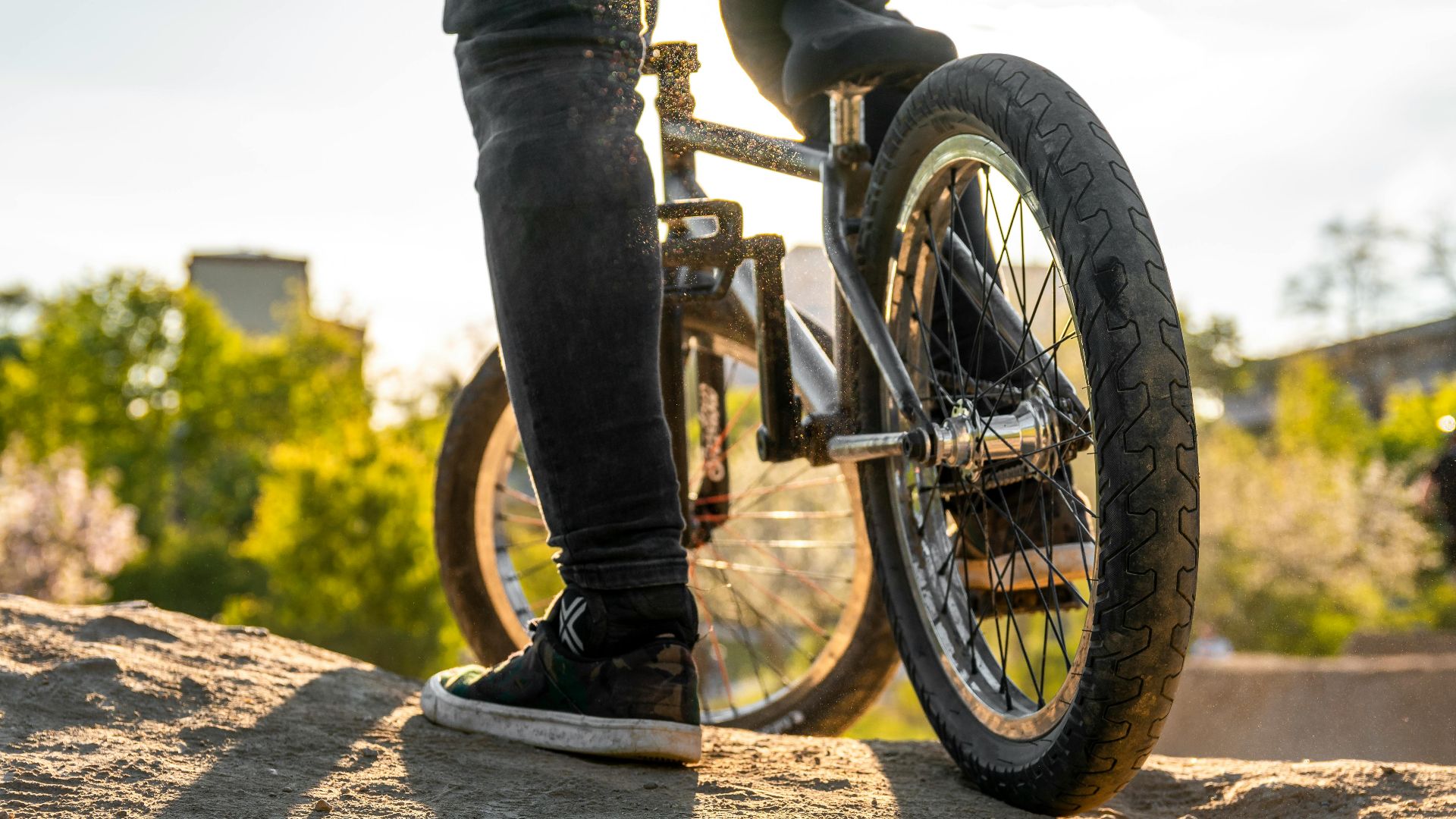 Close-up of a BMX biker standing on dirt in a bright outdoor setting.