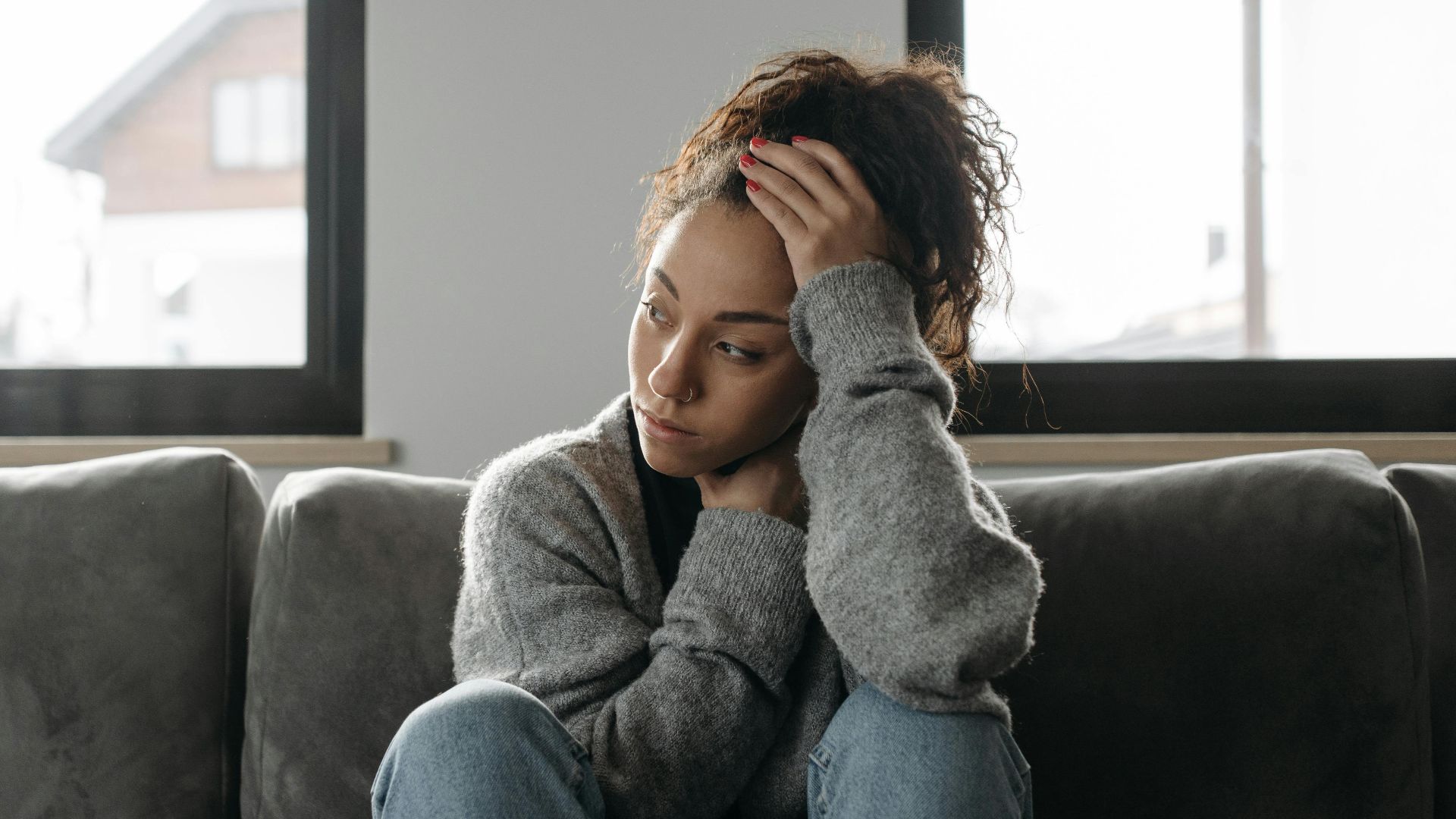 Thoughtful woman in cozy sweater sitting on sofa indoors by the window.