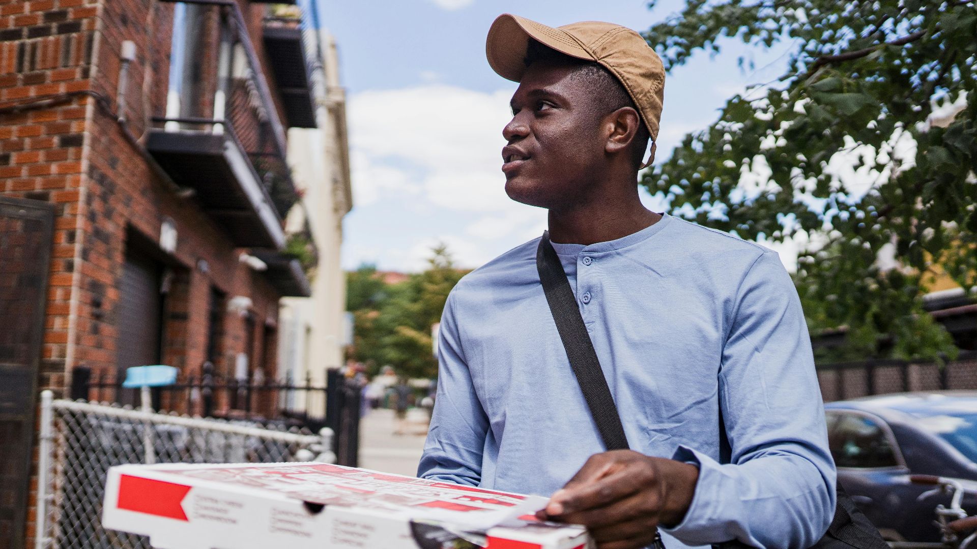 A delivery person with a pizza box walking outside on a sunny day, wearing a cap and blue shirt.