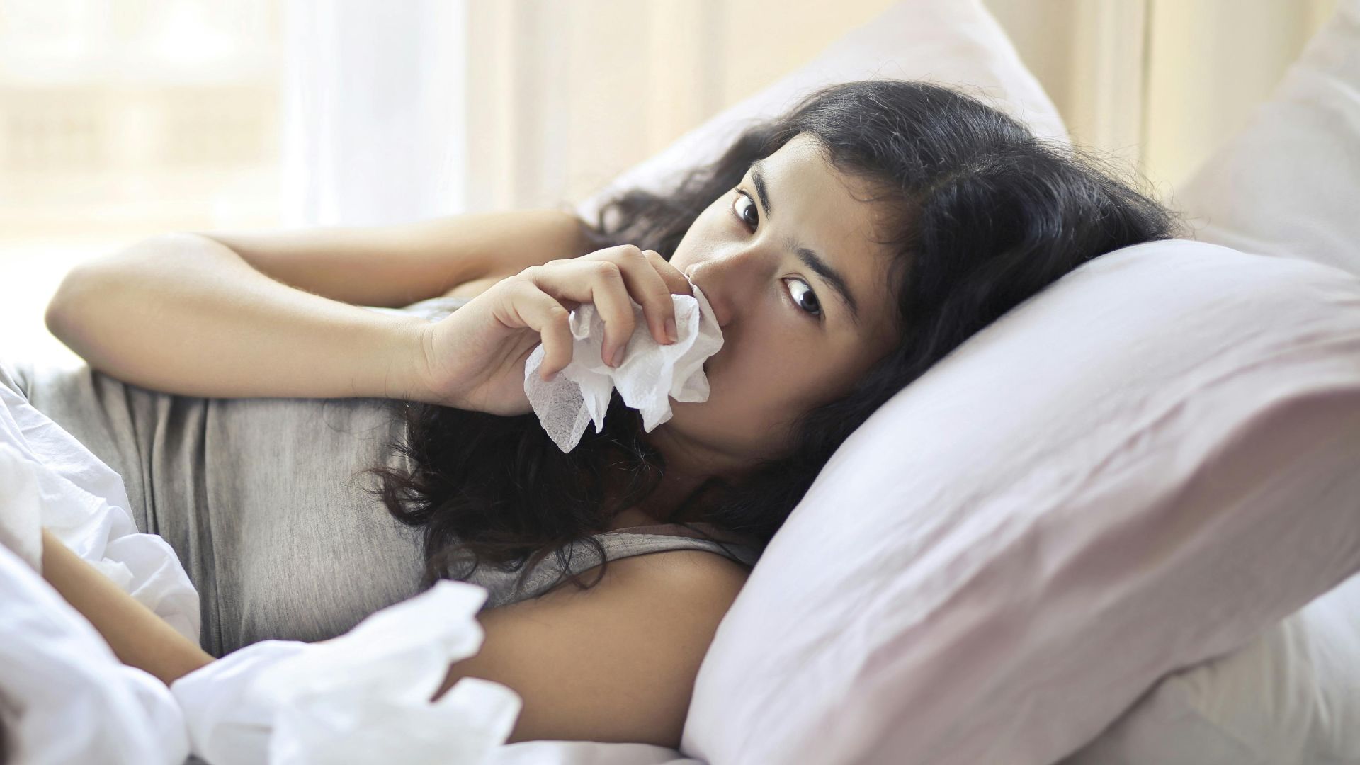 A woman lying in bed indoors, using tissue for cold and flu symptoms.