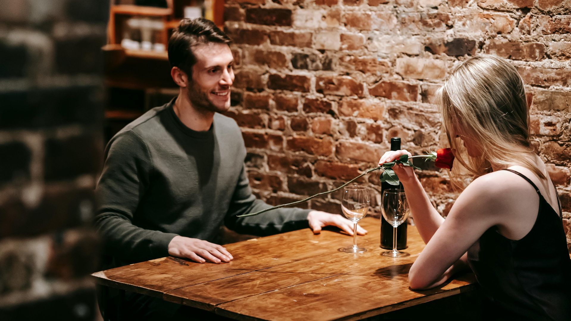 Young couple in elegant clothes sitting in light cafeteria at wooden table near brick wall and having romantic dinner with red rose and wine near glasses
