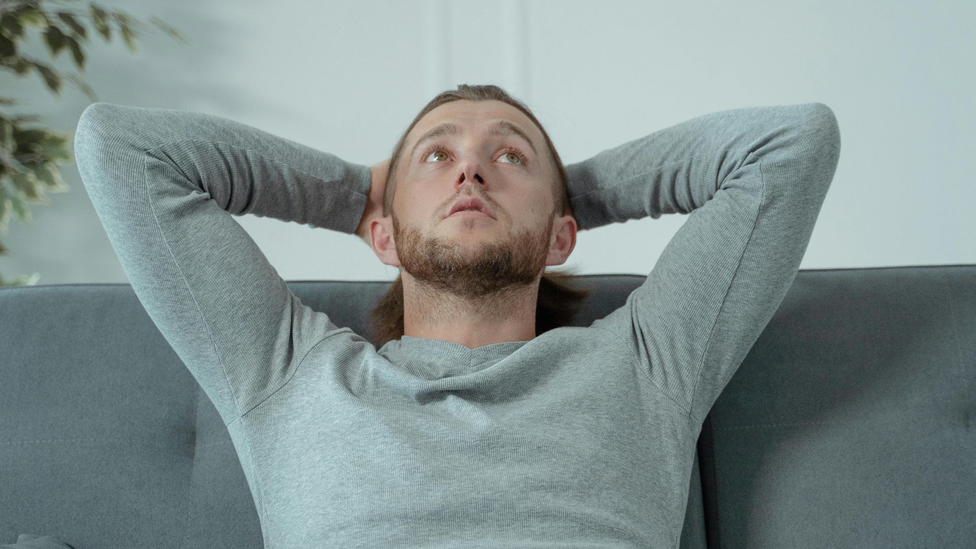 Man looking thoughtful while relaxing on a grey sofa indoors, conveying a sense of contemplation and relaxation.