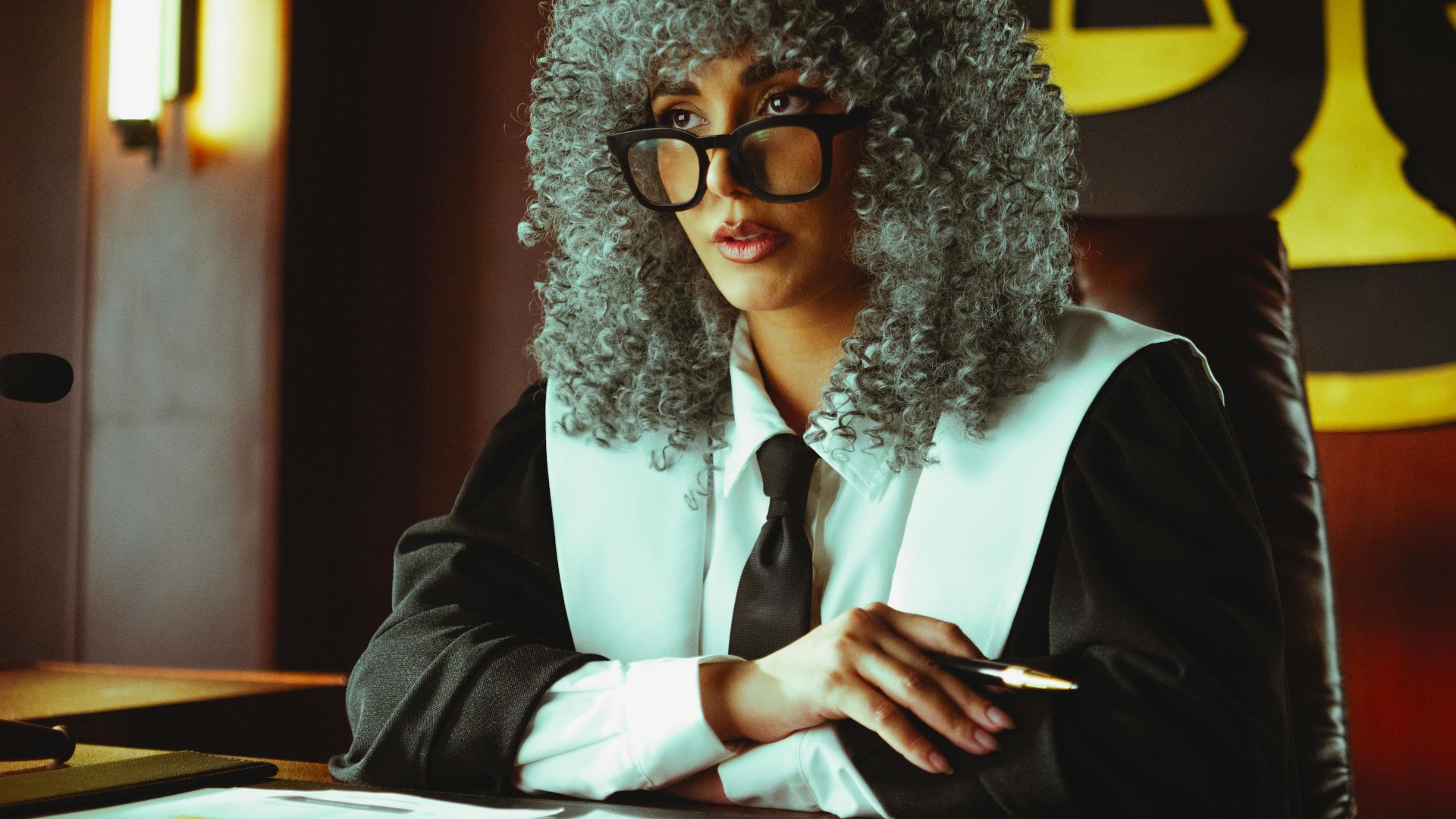 Female judge in courtroom setting, sitting at desk with justice scales in background.