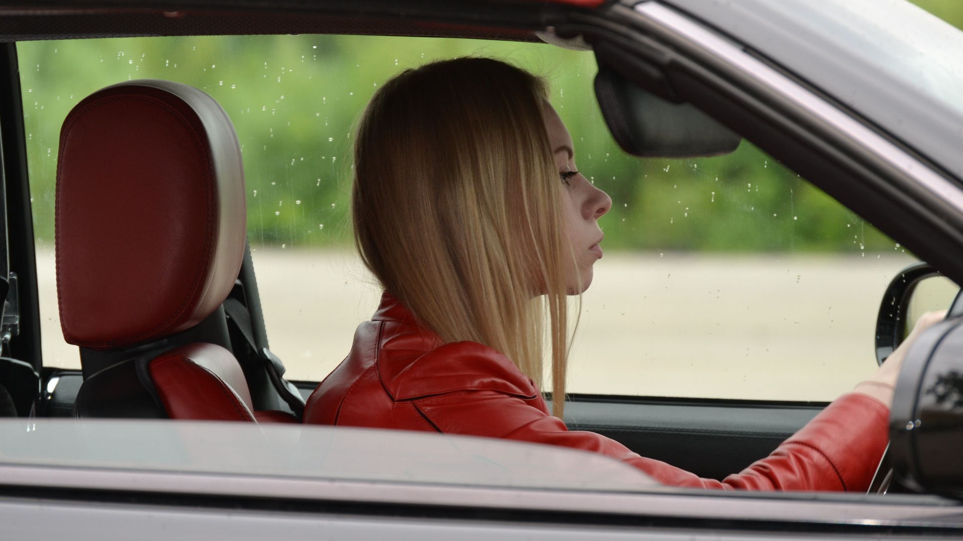 A young woman in a red convertible driving on a rainy day, wearing a red jacket.