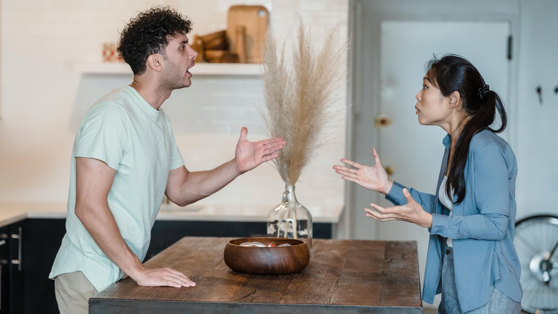A couple engages in a heated argument at a wooden table in a modern indoor setting.