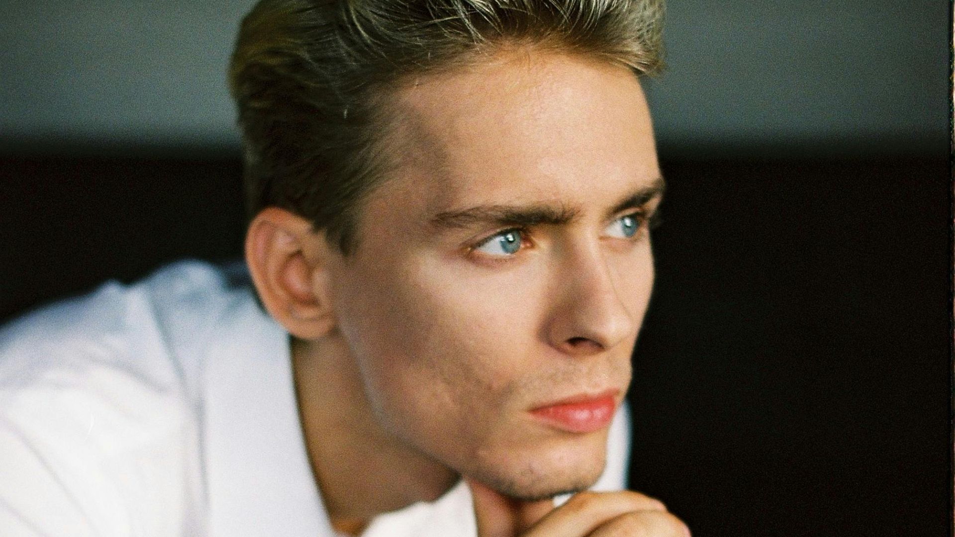 Handsome young guy with blond hair in elegant white shirt touching chin and looking away thoughtfully while sitting on chair in room