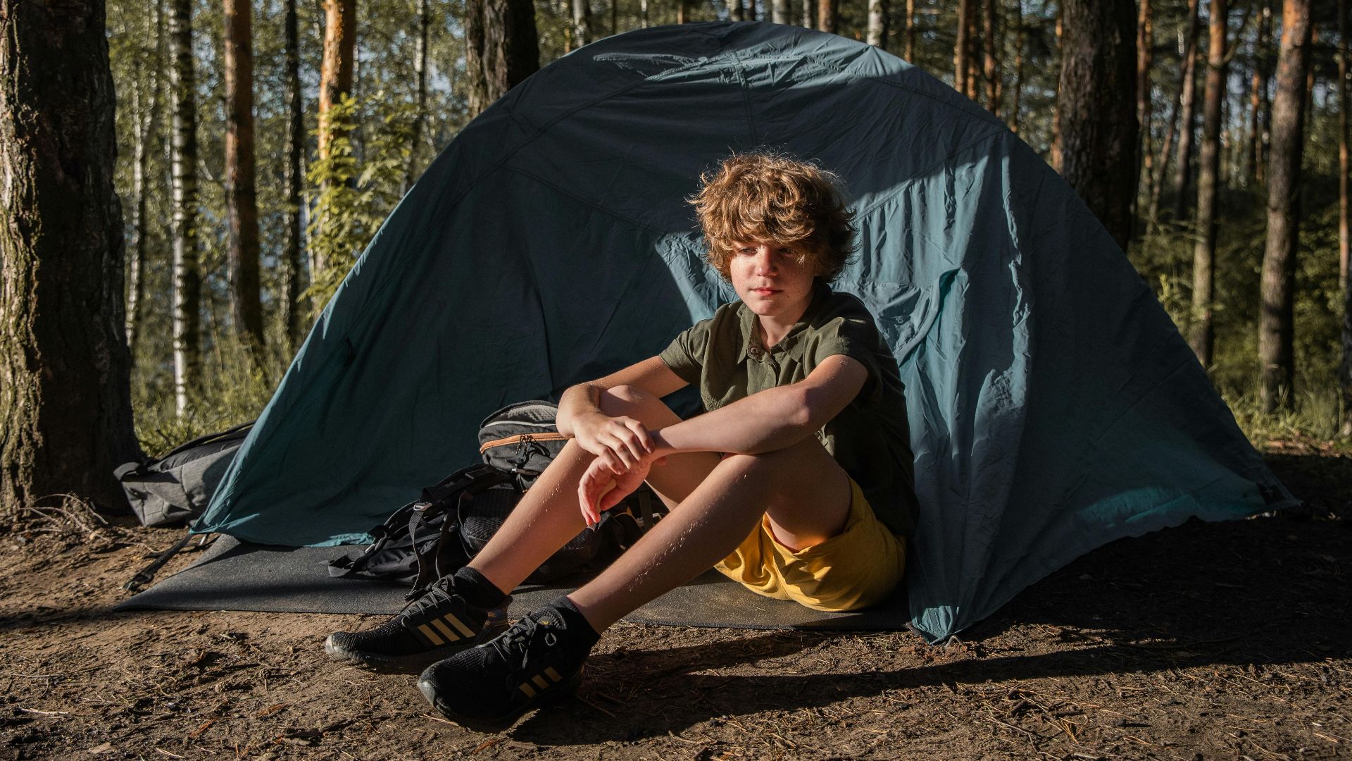 A young boy enjoying a camping trip in a forest, sitting in front of a tent.