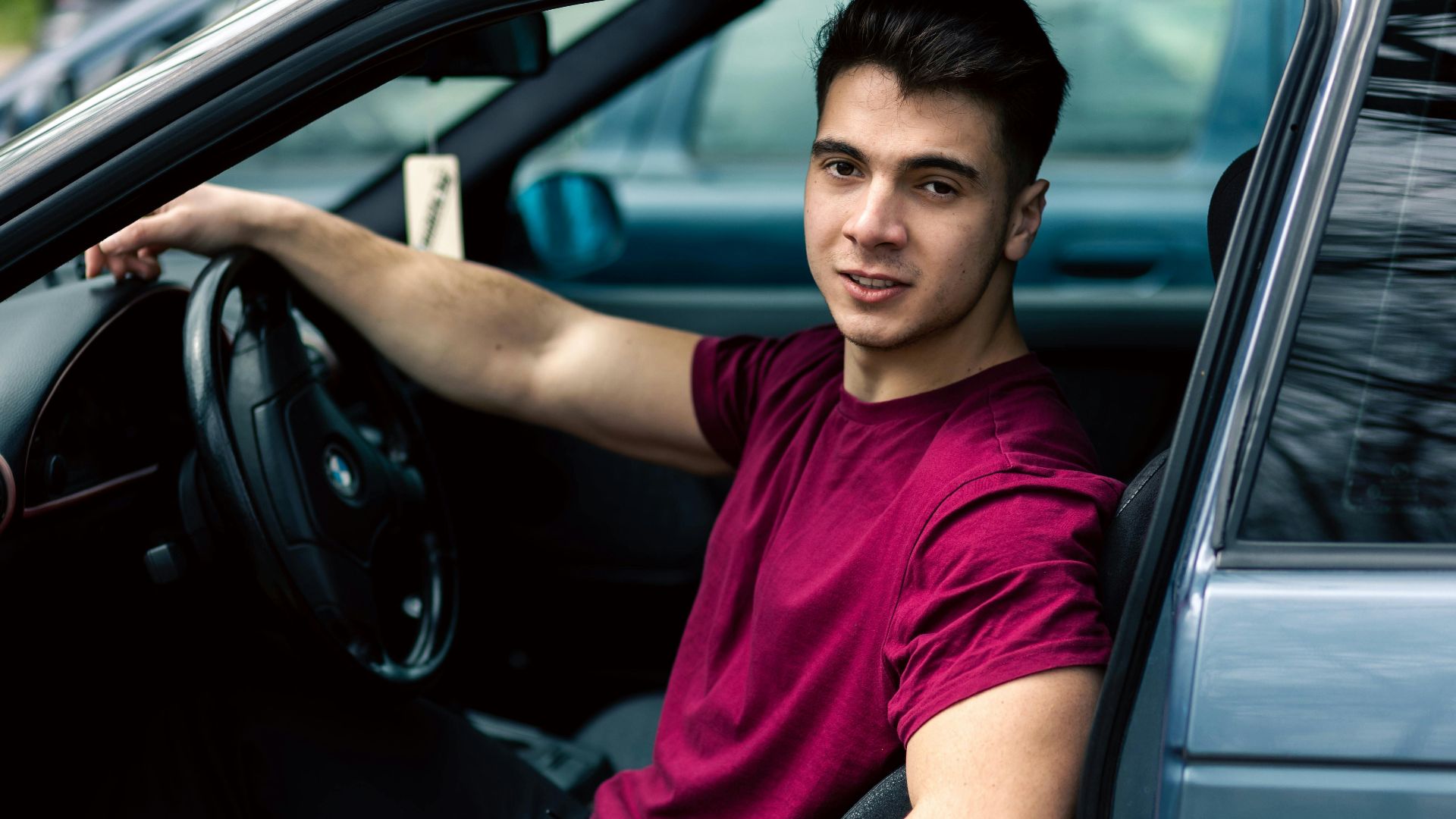 A young man wearing a maroon t-shirt sits casually in a blue car, portraying a relaxed and modern lifestyle.