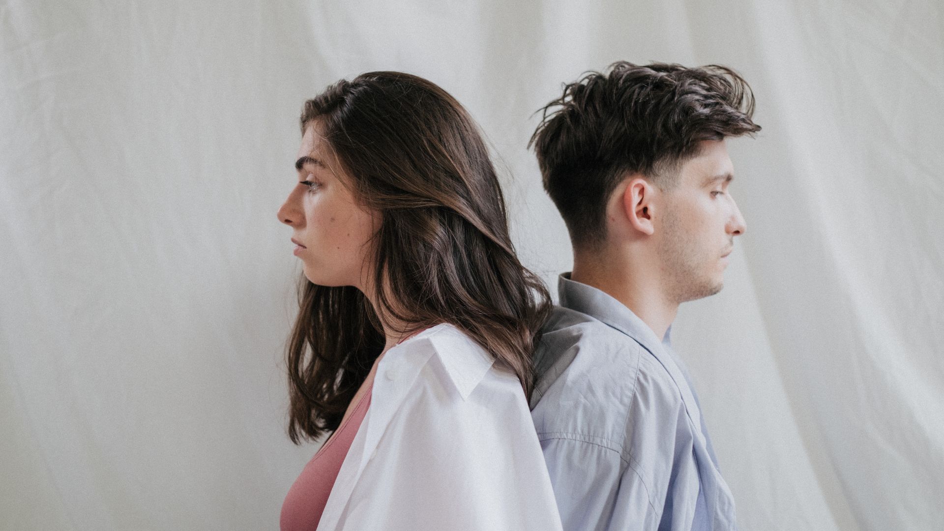 A man and woman sit back to back in a studio setting, expressing contemplation.