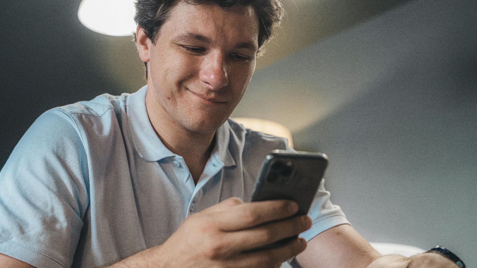 A man in a blue polo shirt smiling while using a smartphone indoors.