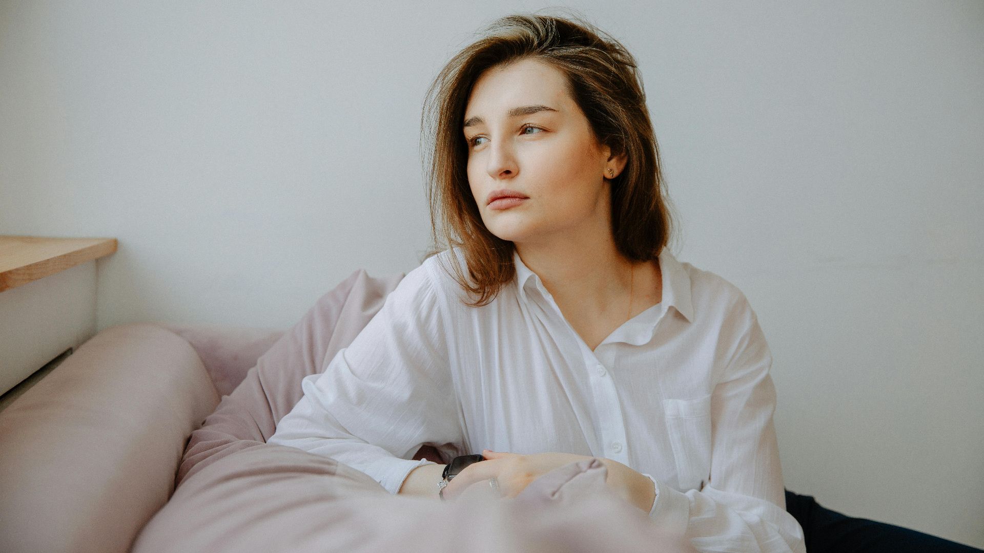 A contemplative woman in a white blouse sitting on a sofa, looking away.