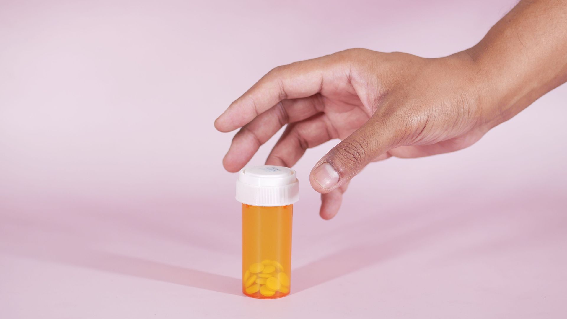 A hand reaches for a prescription bottle with yellow pills on a pink background, symbolizing healthcare.