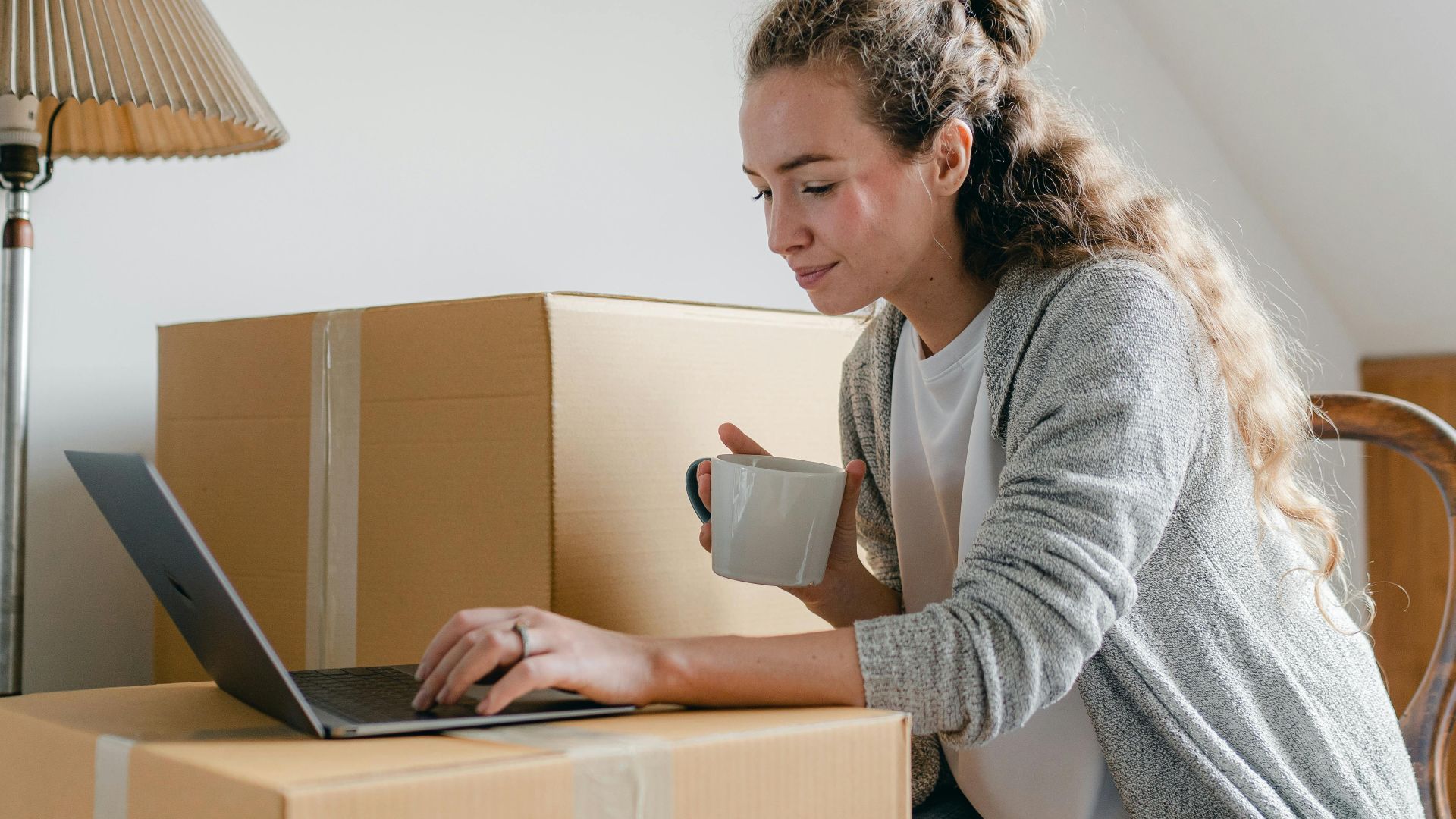 Content lady in casual wear typing on netbook while sitting on chair with cup of hot drink near big cardboard boxes in light apartment