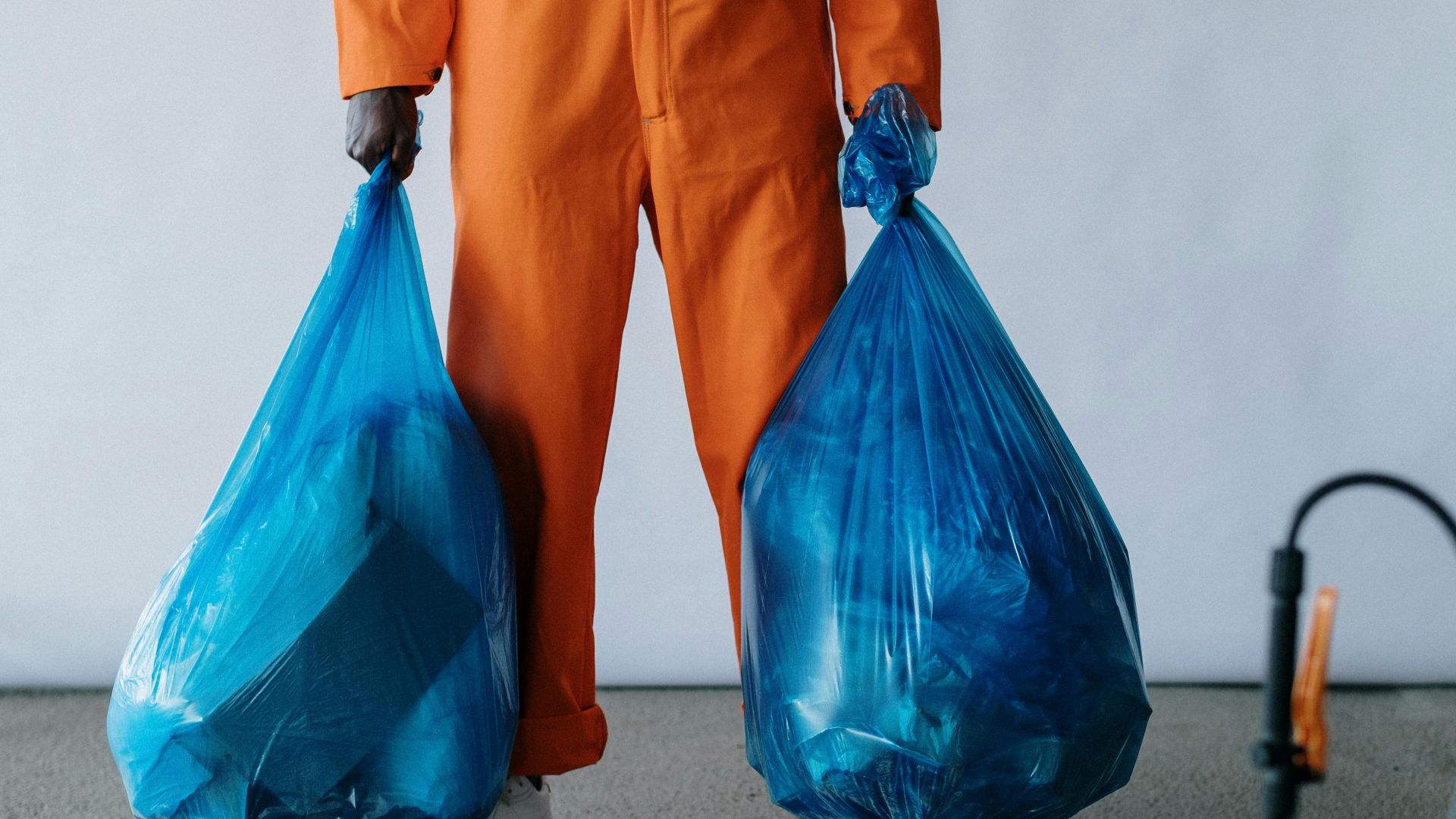 Sanitation worker in orange overalls holding blue trash bags indoors for waste management.