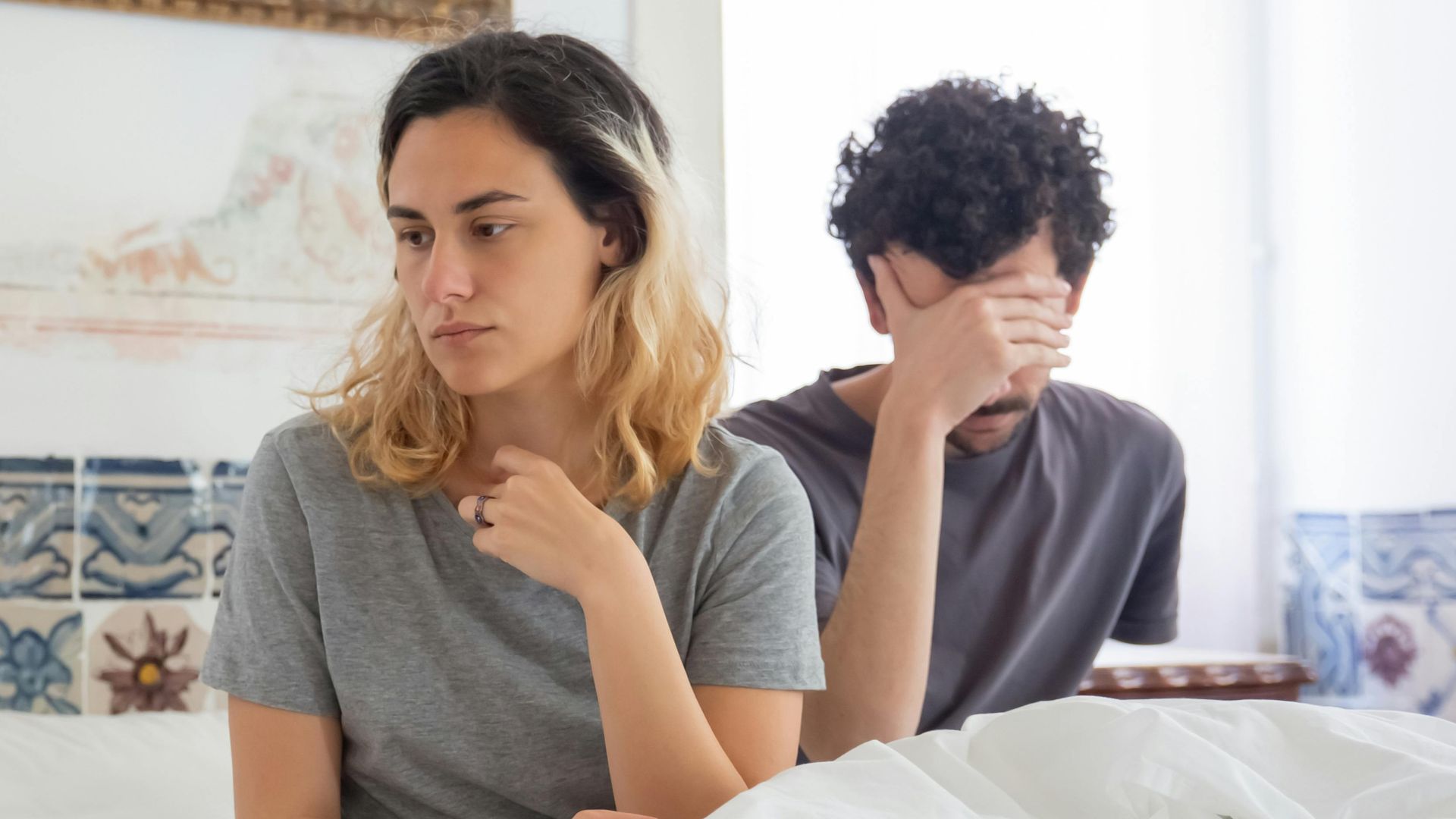 A couple sits together in a bedroom, experiencing a moment of misunderstanding.