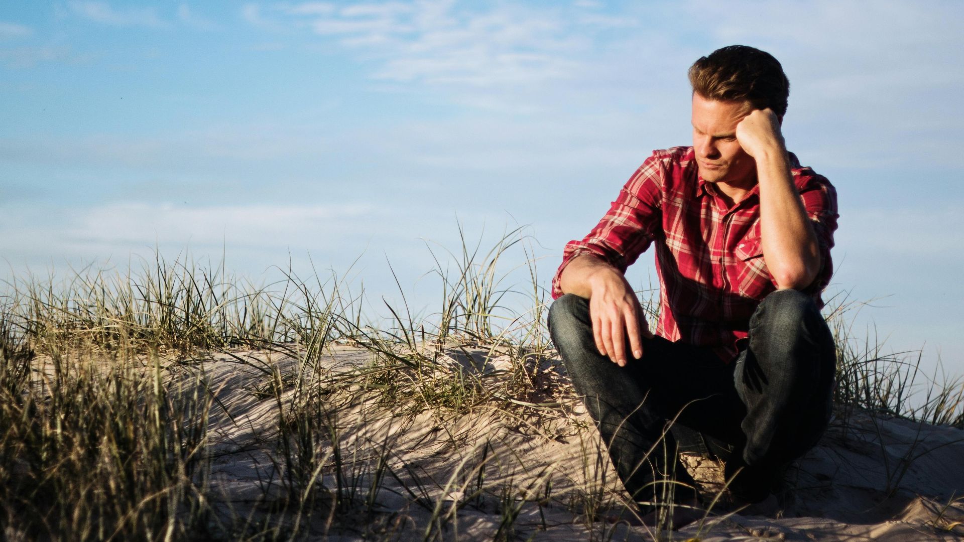 Man in a red plaid shirt sitting thoughtfully on a sandy dune in Australia, surrounded by grass.