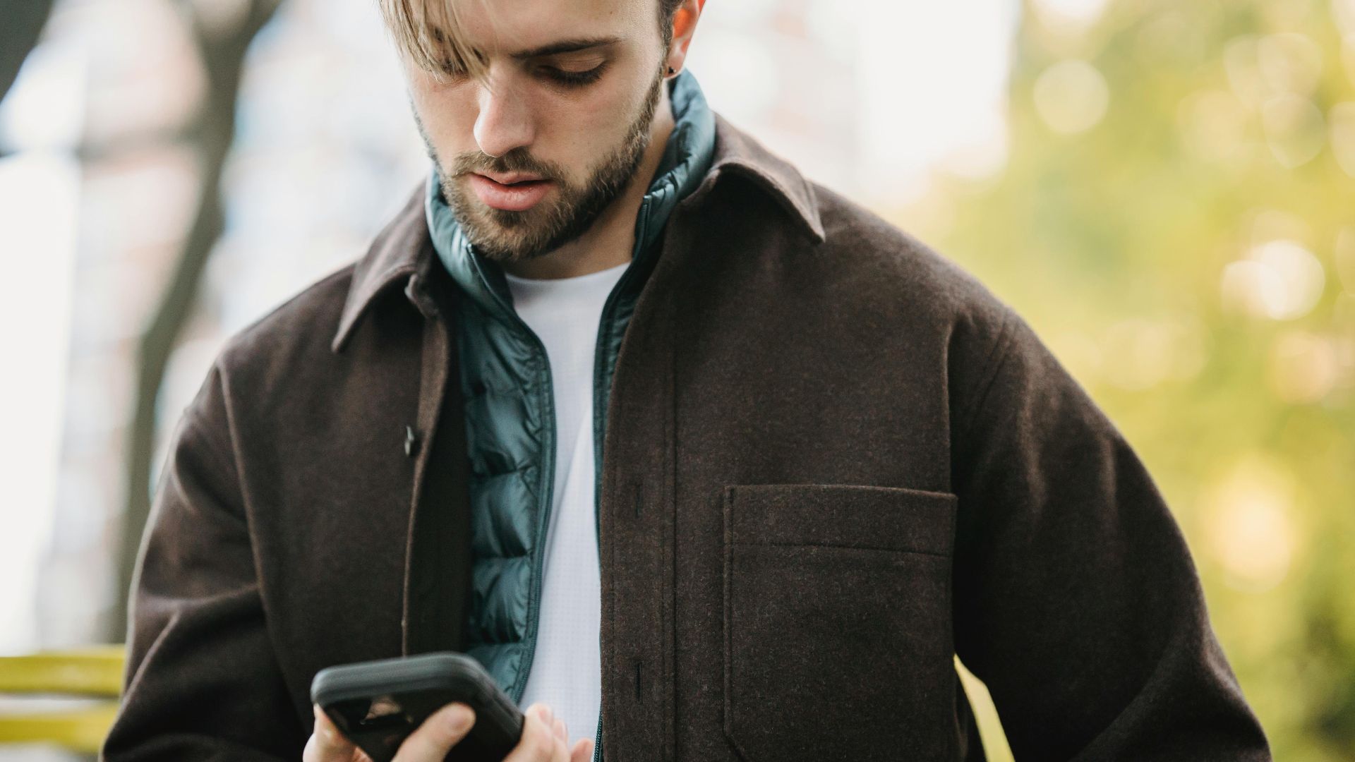 Young bearded man in casual clothes receiving text message on smartphone while standing on street