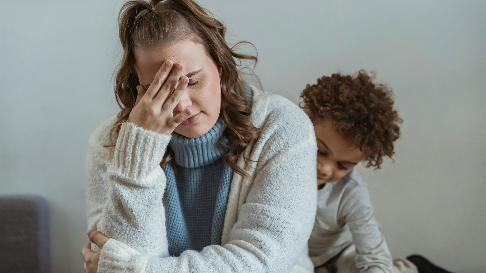 Frustrated mother with hand on forehead and closed eyes sitting near African American son near wall in room at home