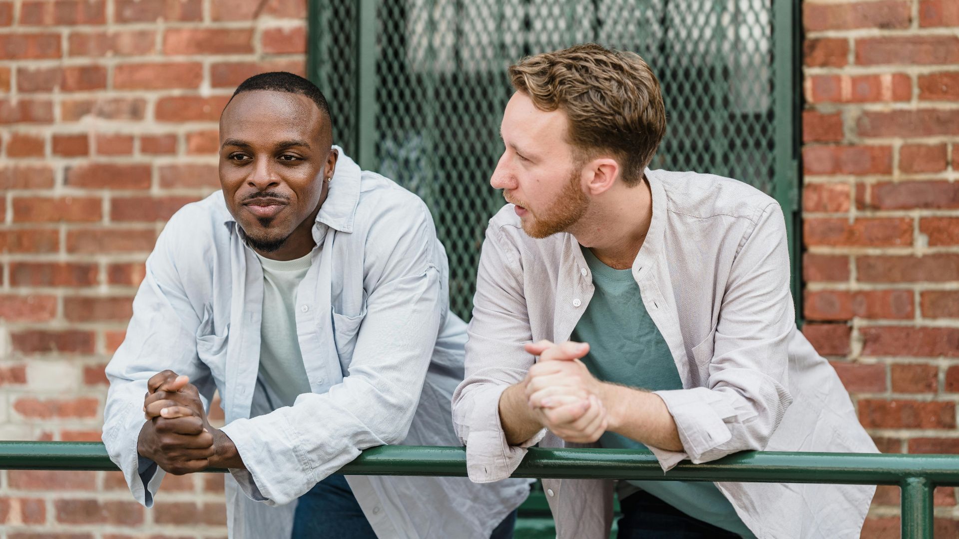 Two men having a relaxed conversation outside against a brick wall backdrop.