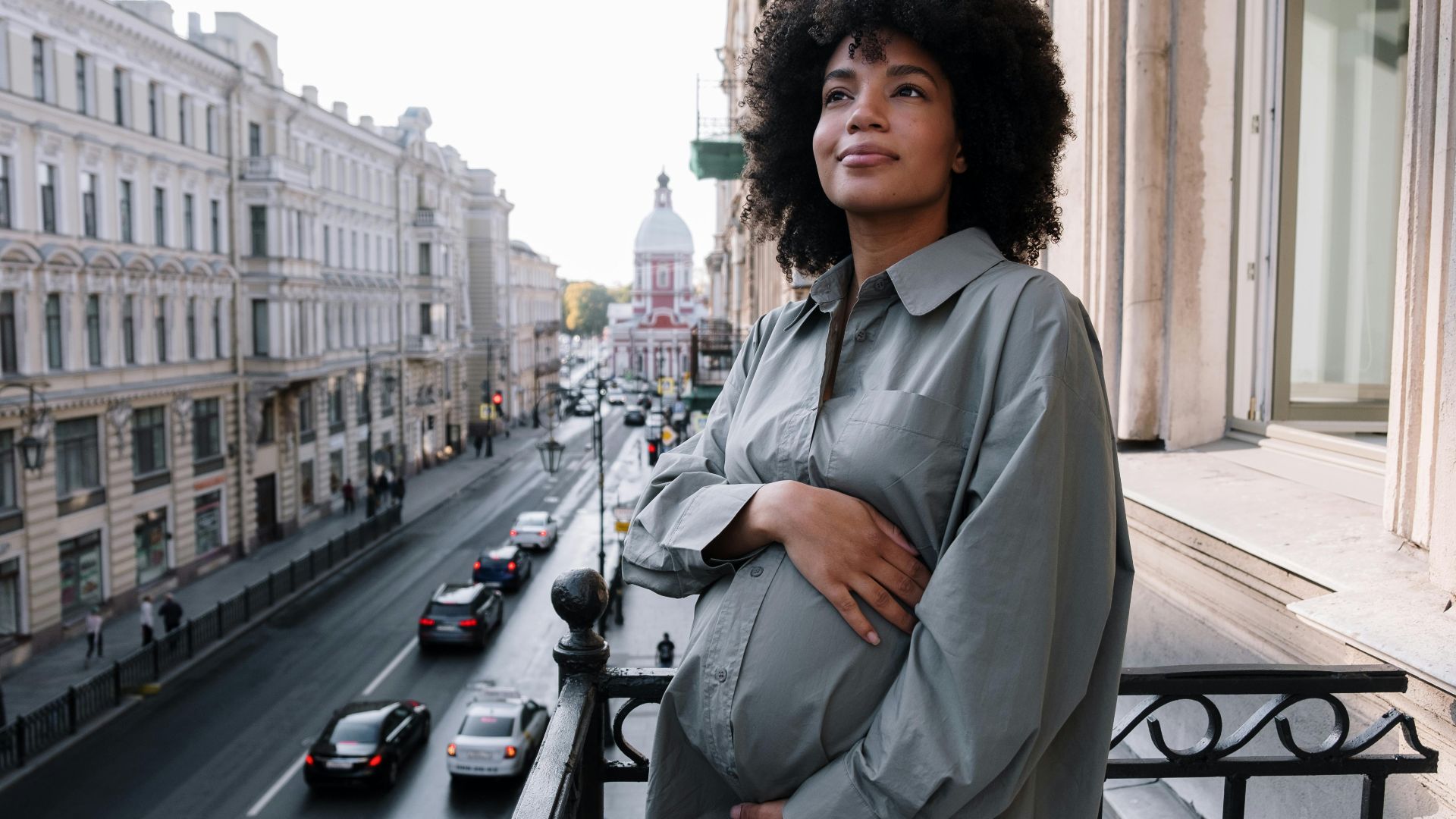 A pregnant woman stands on a balcony overlooking a bustling city street, radiating tranquility.