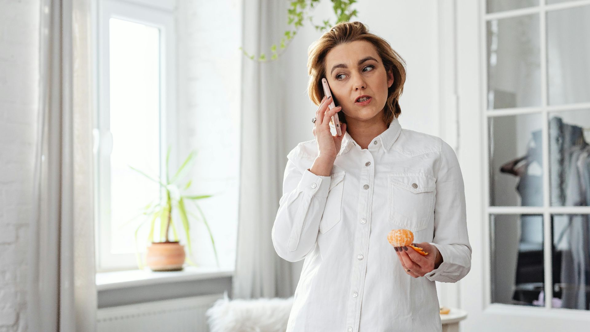 Woman in casual attire talking on phone while eating, in home office setting.
