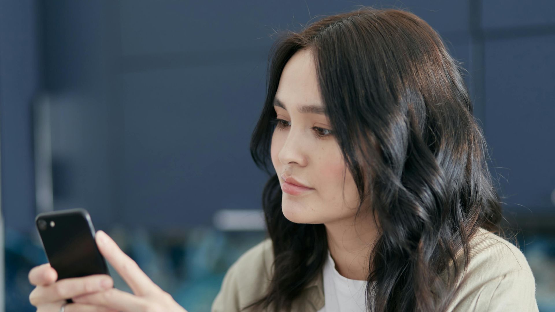 A young woman with black hair using a smartphone indoors, focused on the screen.