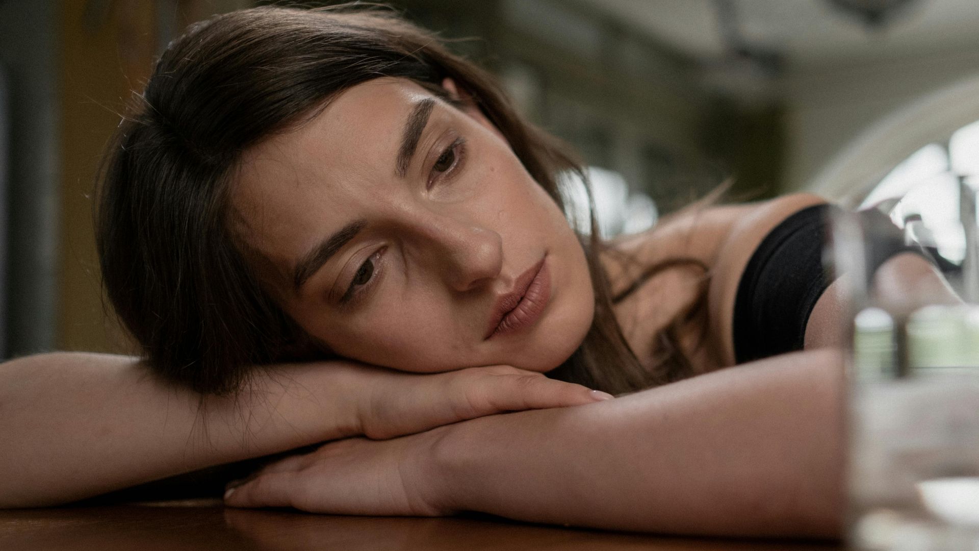 Pensive woman resting her head on a wooden table with a glass of water nearby, indoors.