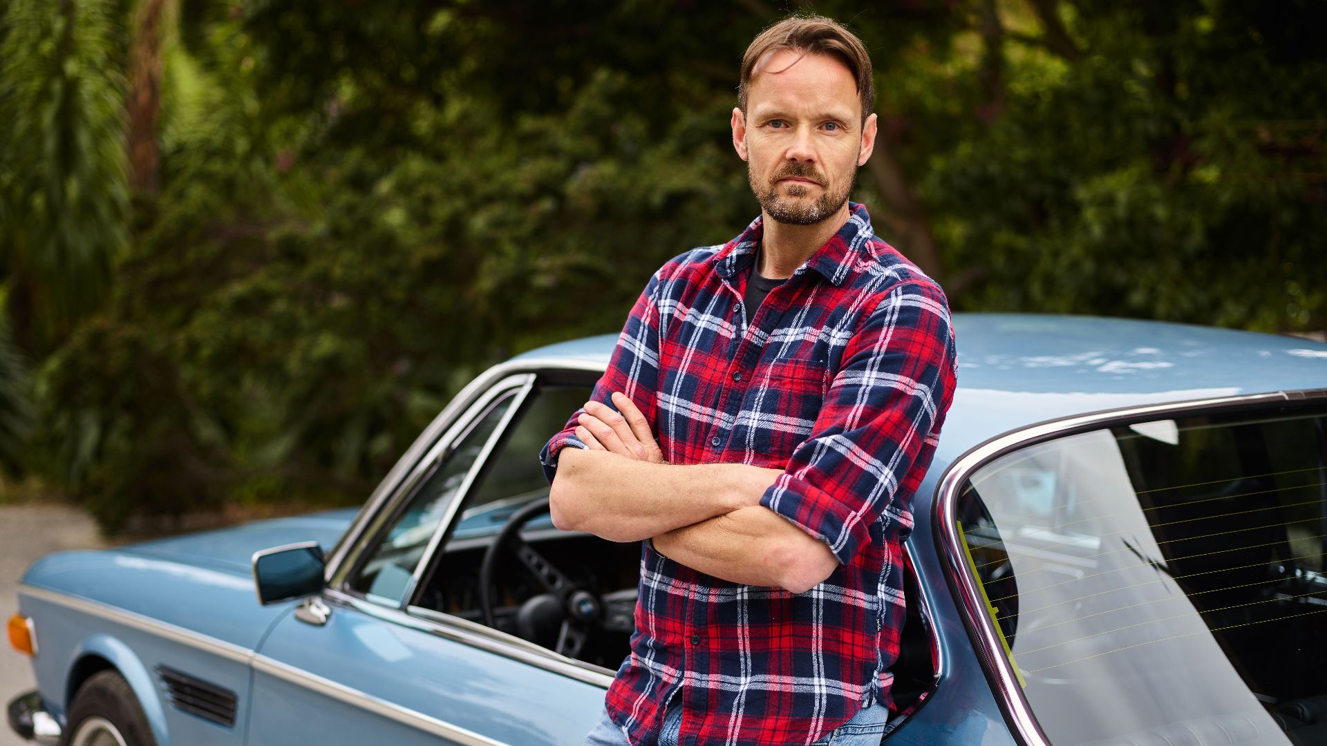 Adult bearded male in jeans and shirt standing with crossed arms leaned on blue retro auto near forest in daylight and looking at camera