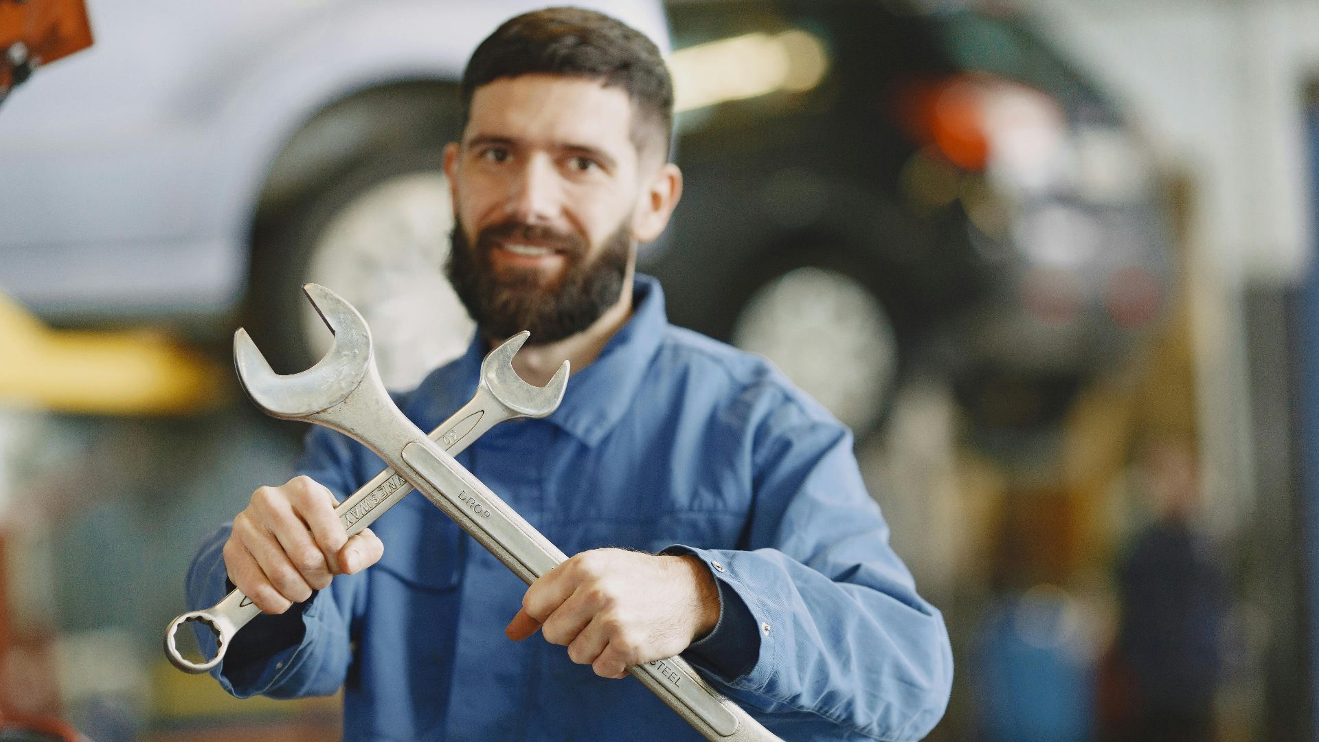 Mechanic in blue uniform holding wrenches inside an auto repair shop, ready for vehicle maintenance.
