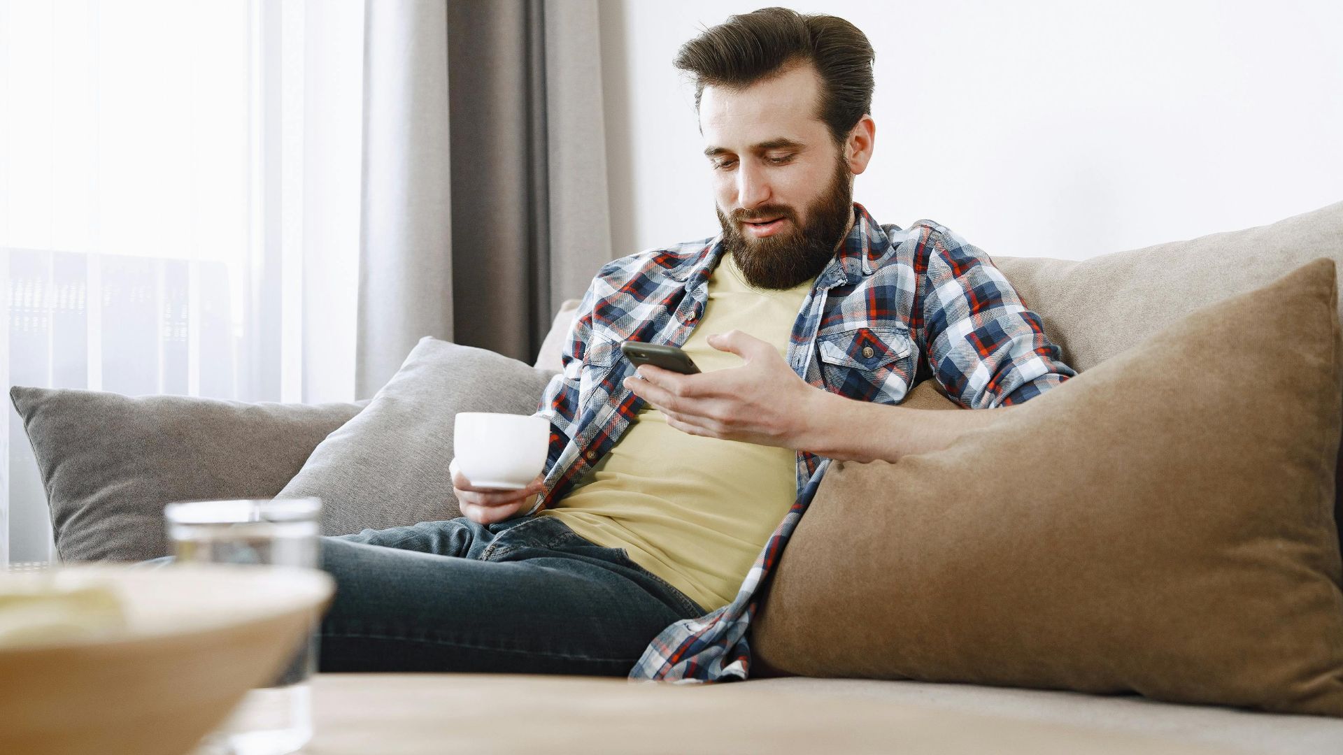 A man sits comfortably on a sofa using his smartphone while enjoying a cup of coffee.