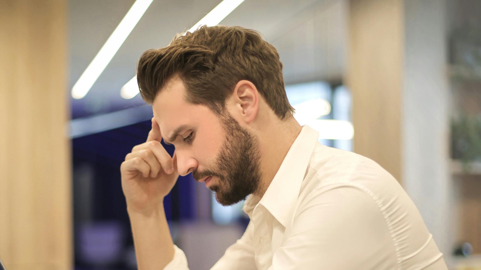A thoughtful man in a shirt works on his laptop at a modern indoor office space.