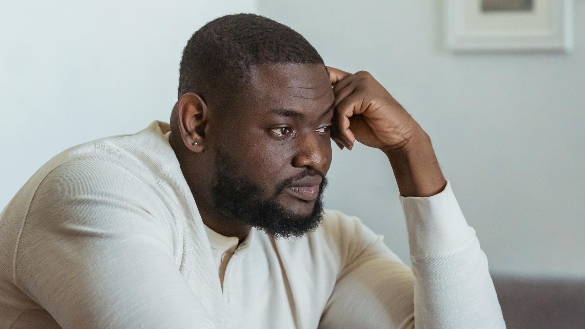 Depressed African American sitting on soft couch near table with papers and laptop and pondering