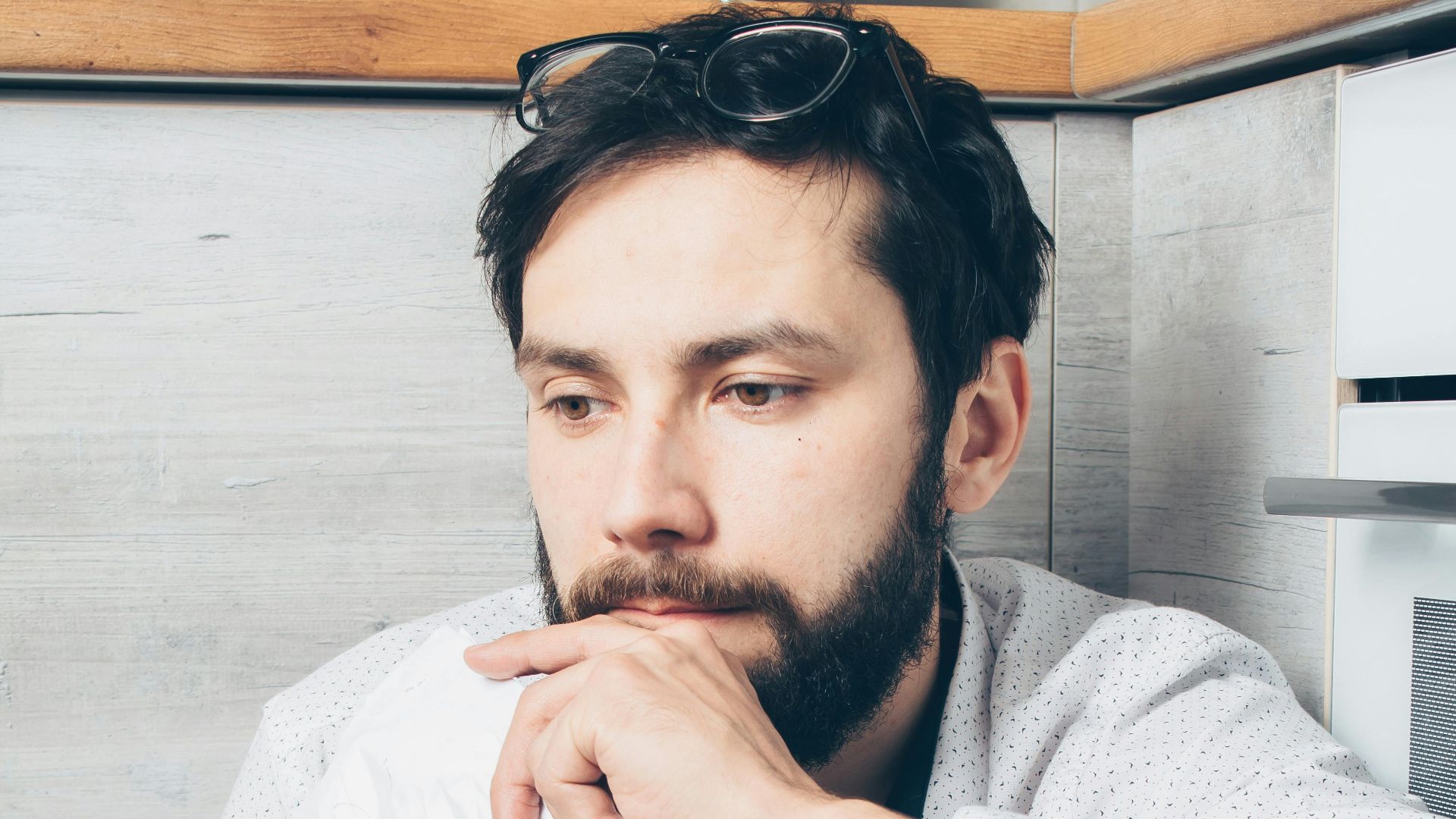 A young bearded man in deep thought holding a tissue, sitting indoors.