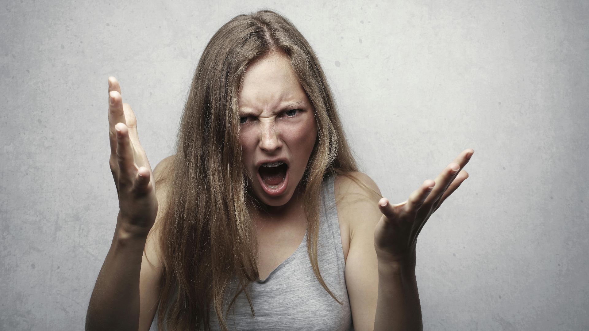 A young woman showing intense anger and frustration, yelling with hands raised against a gray background.