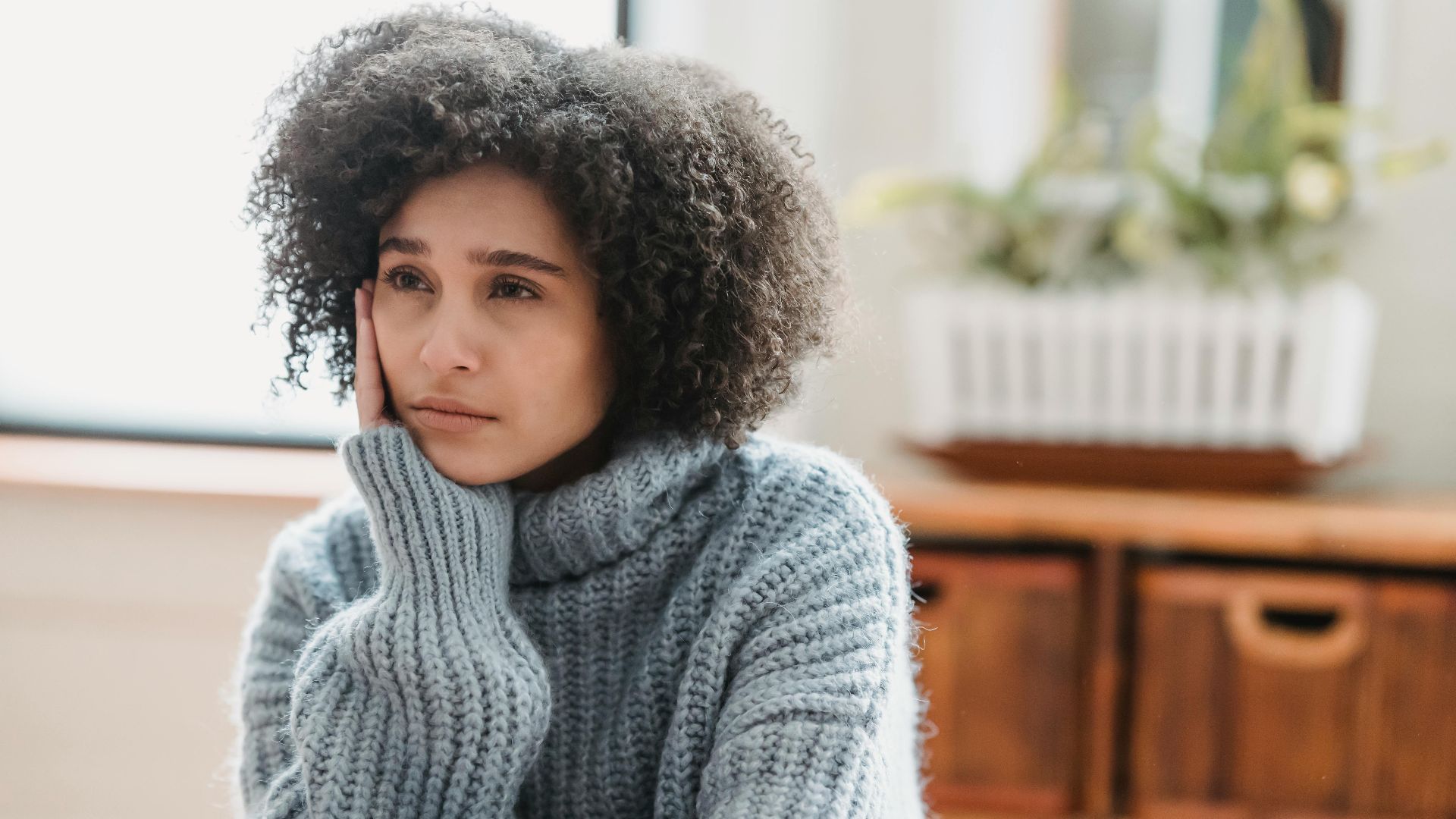 Discontent African American female leaning on hand and looking away while sitting in light room near green plant on blurred background