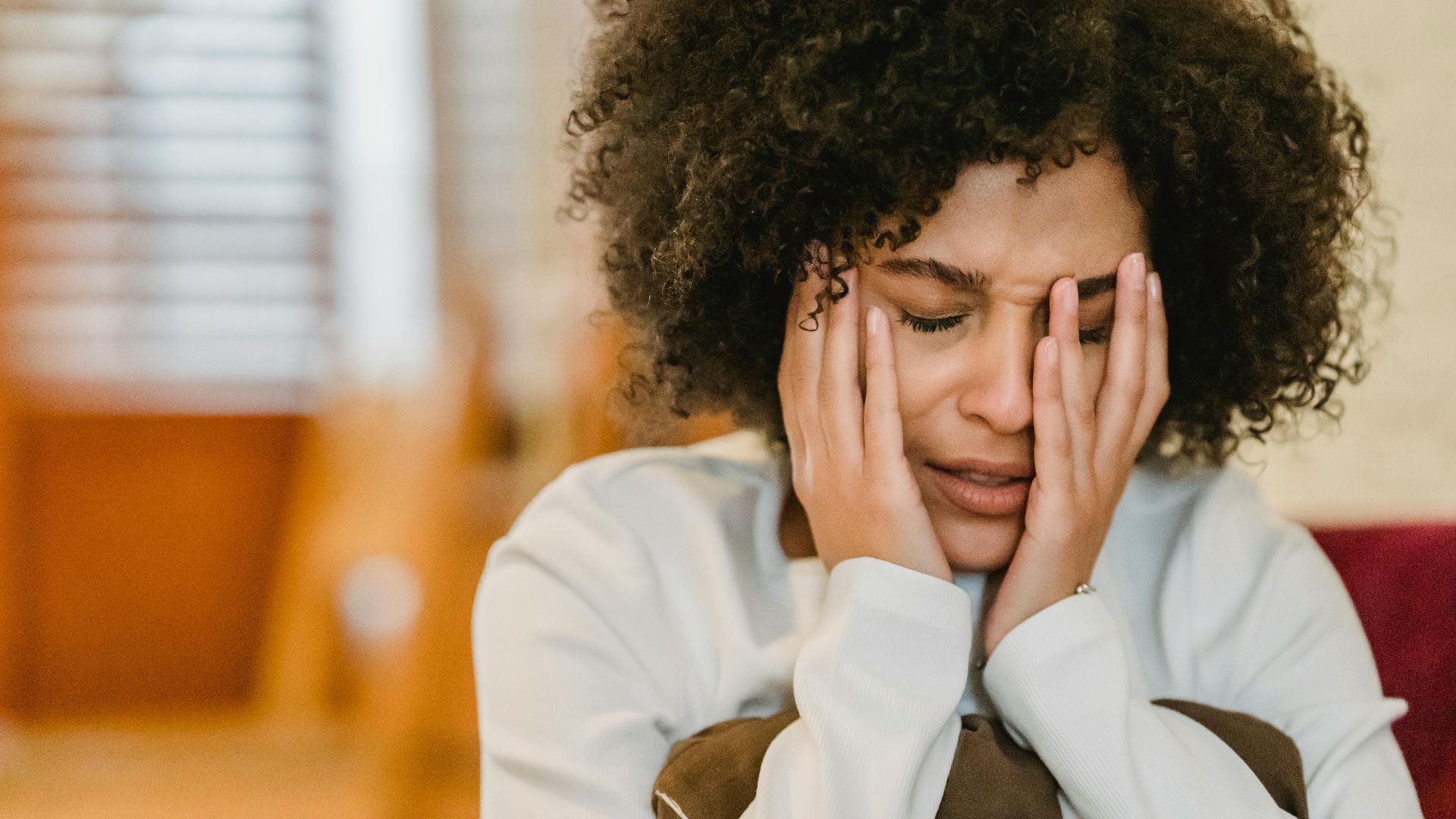 Despaired African American female with closed eyes touching face while sitting with pillow in light room at home on blurred background